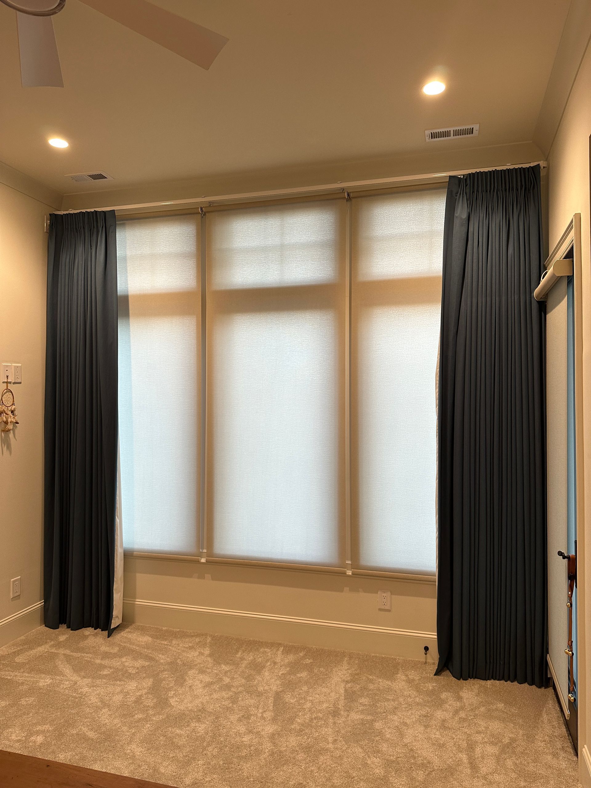 Dark curtains frame a triple-window with light-colored shades. The room has light walls and carpet.