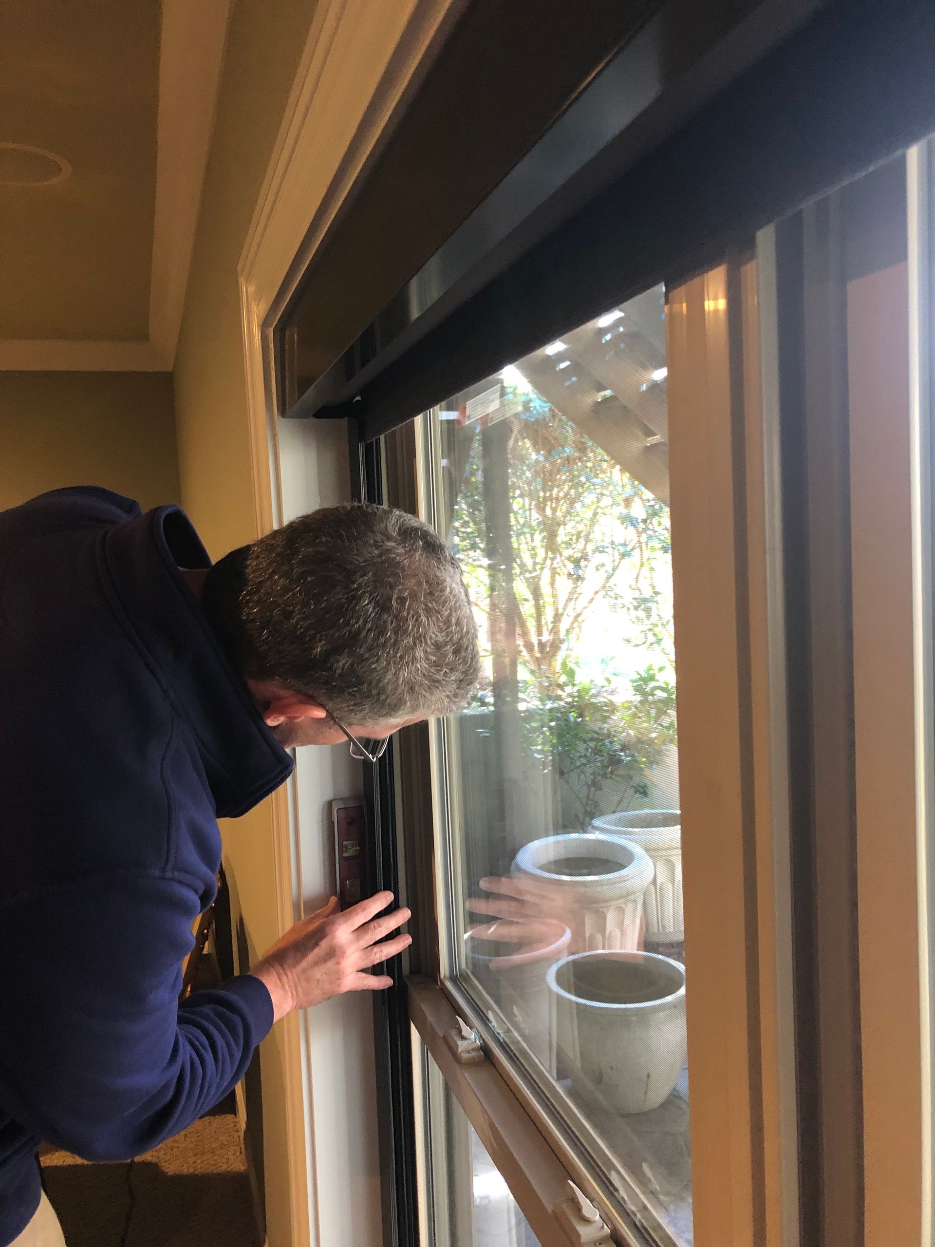 Person examines a window frame, indoors. The window overlooks a yard with potted plants.