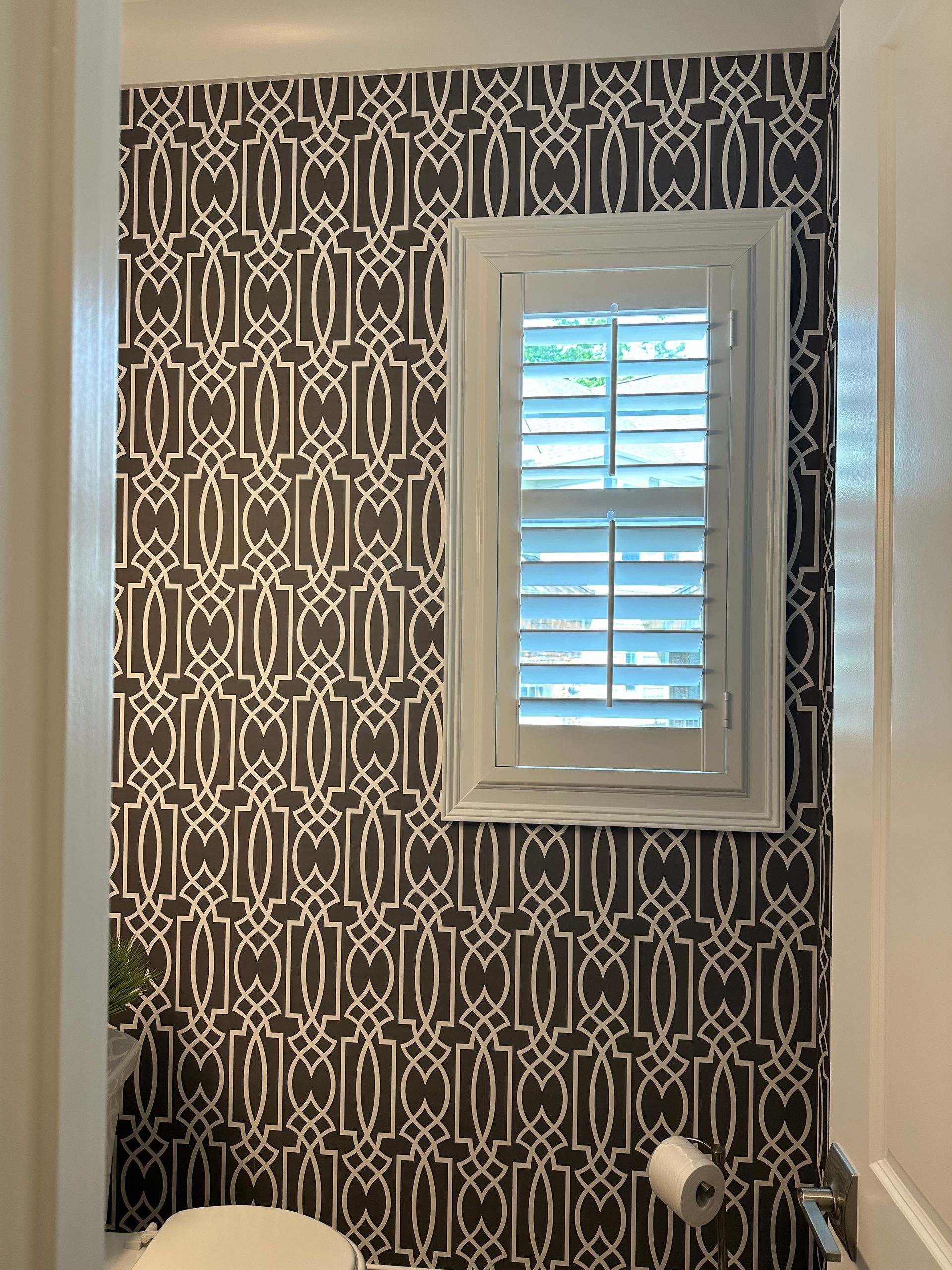 Dark geometric patterned wallpaper in a small bathroom with a window and white shutters.