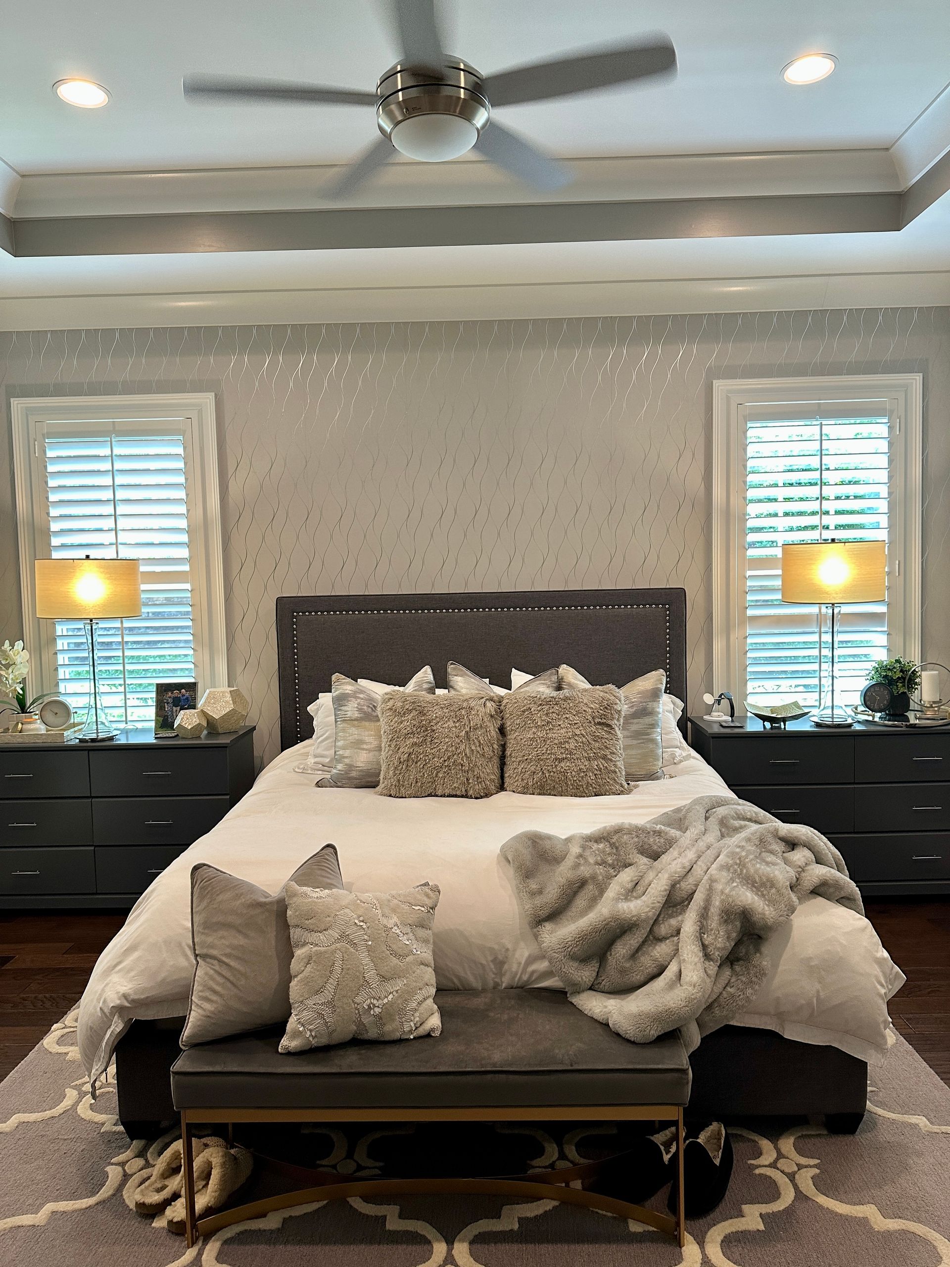 Bedroom with bed, nightstands, windows, and ceiling fan. Gray and white color scheme, plush bedding.