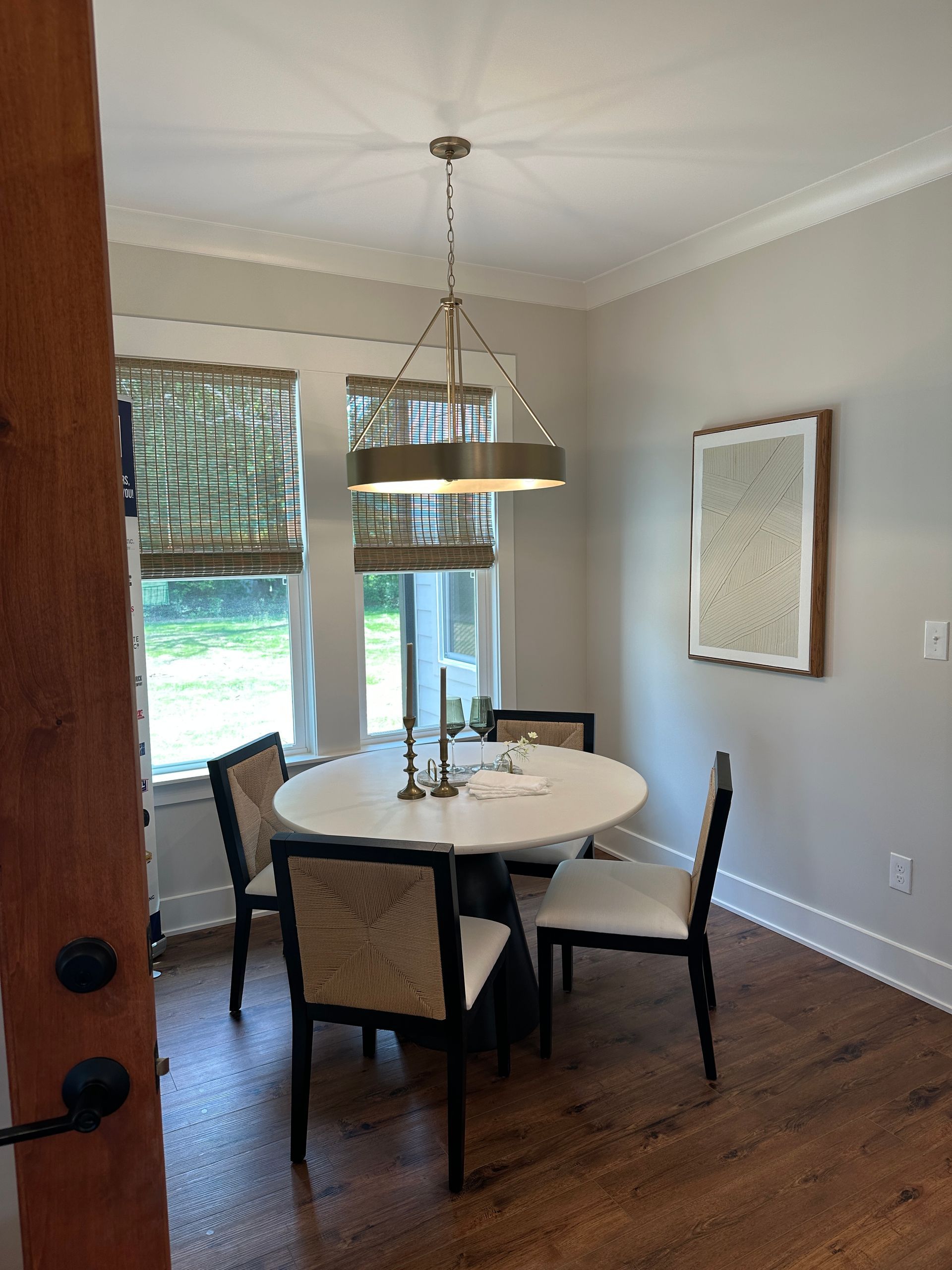 Dining room with a round table, four chairs, and a chandelier. Windows with patterned shades and a framed artwork.