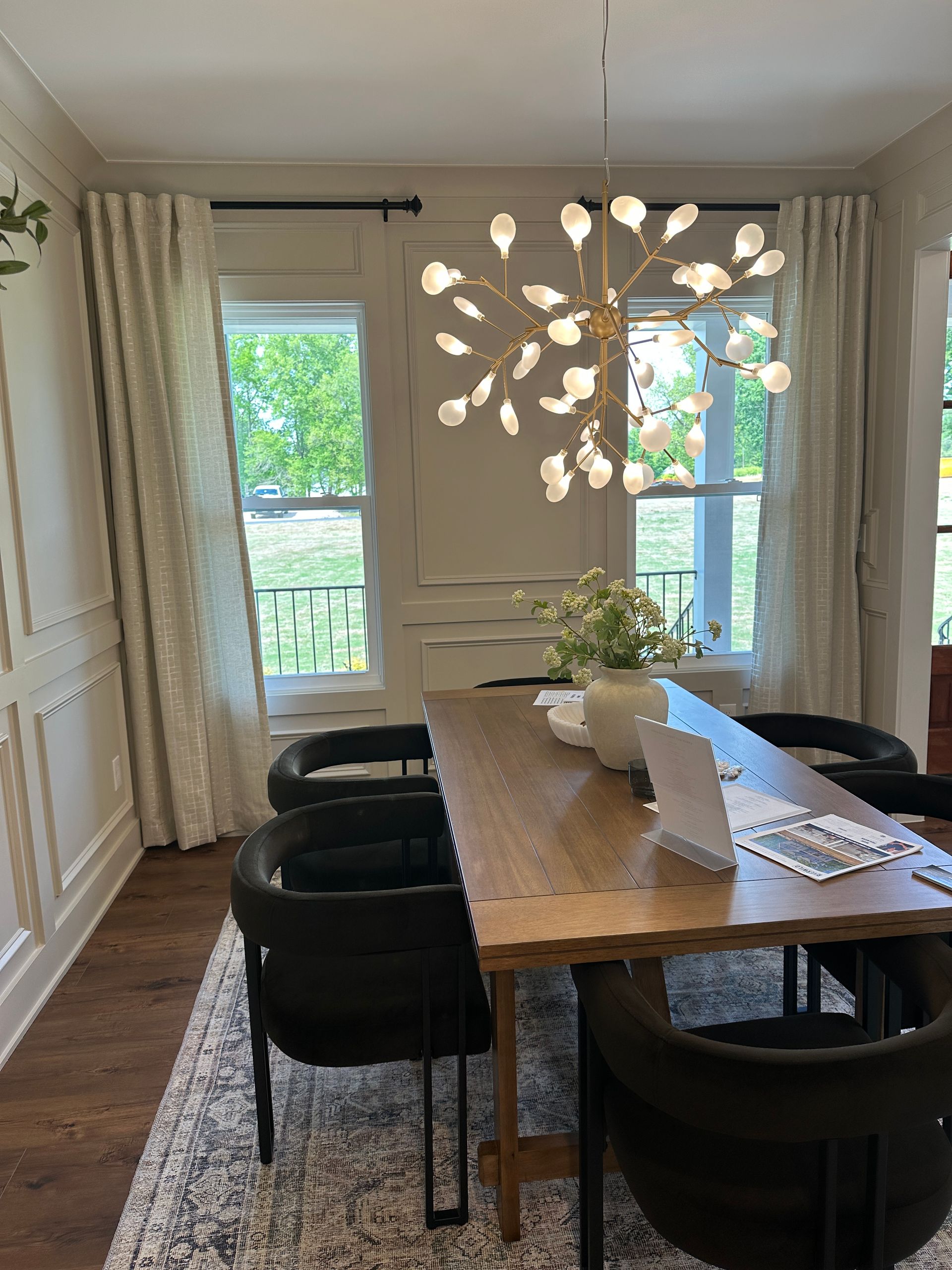 Dining room with wooden table, black chairs, and floral chandelier, curtains on windows.