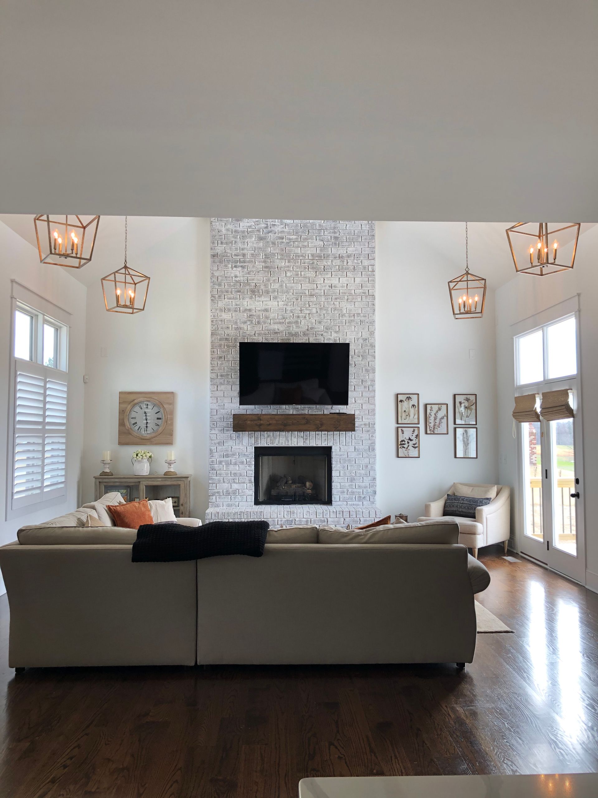 Living room with white brick fireplace, TV, and cream-colored furniture. Hardwood floors and tall windows.