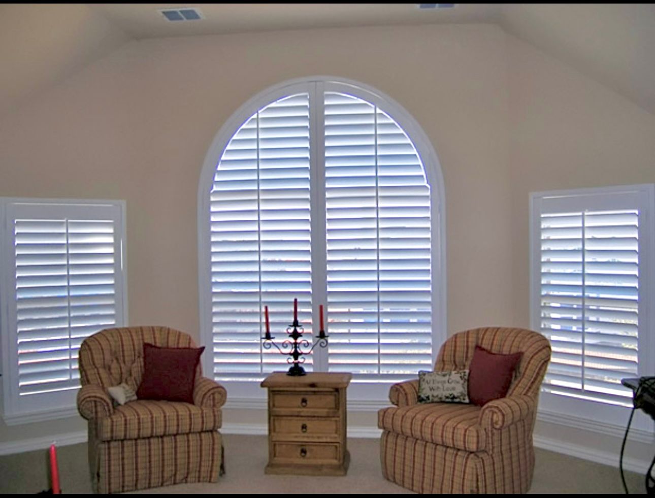 Living room with arched window and shutters, two armchairs, side table, and candlesticks.
