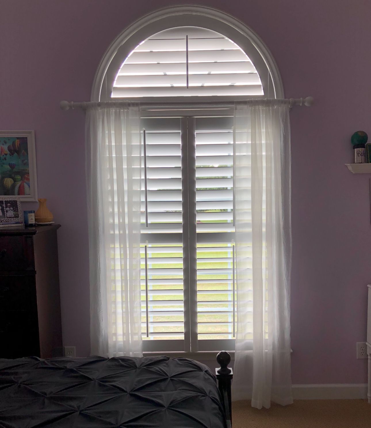 Window with white shutters and sheer curtains, in a room with a lilac-colored wall and dark bed.