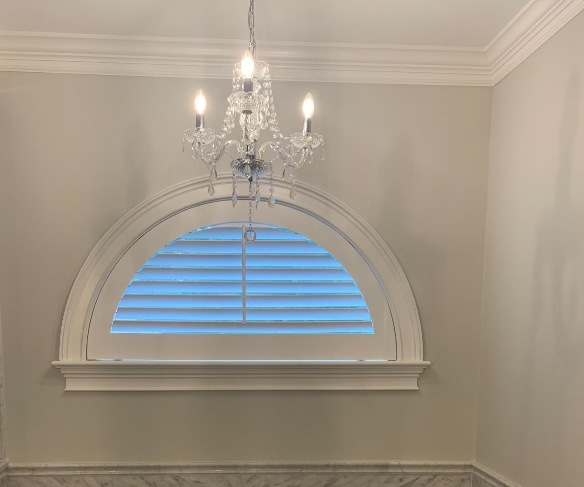 A chandelier hangs above a half-moon window with white shutters. Pale gray walls and crown molding surround.