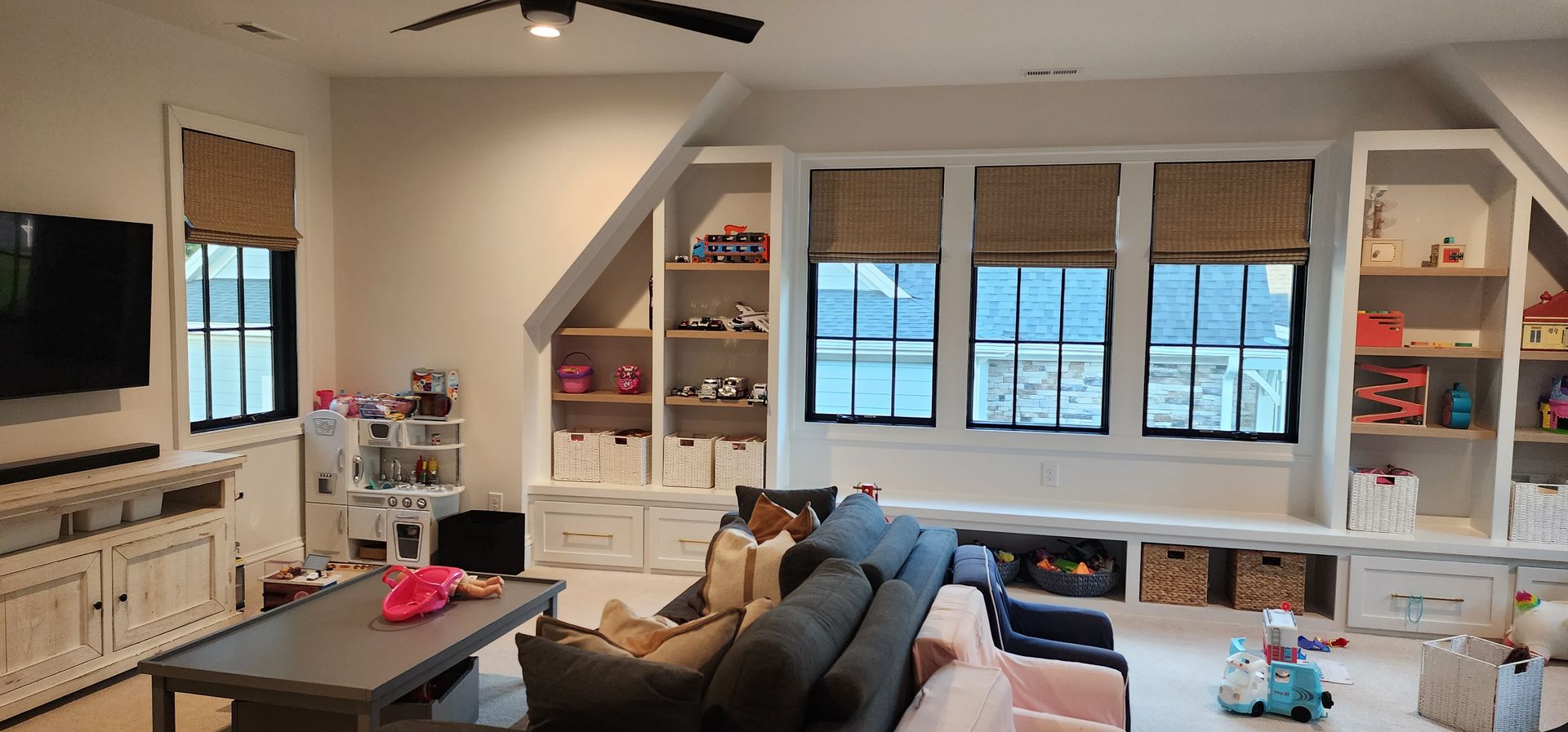 Playroom with built-in storage, a gray couch, a coffee table, and windows with woven shades.