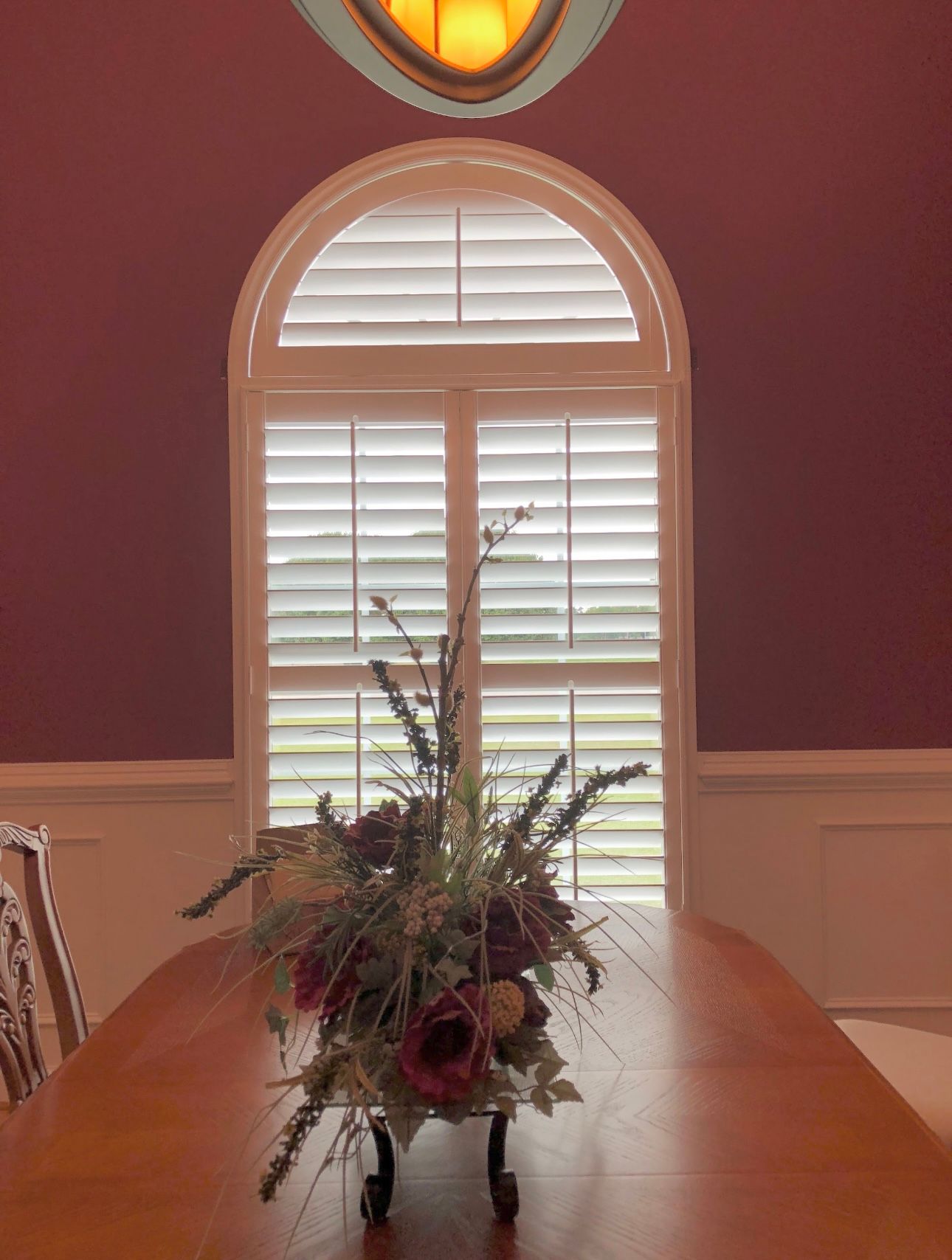 Table set with floral arrangement in front of an arched window with closed white shutters; burgundy walls.