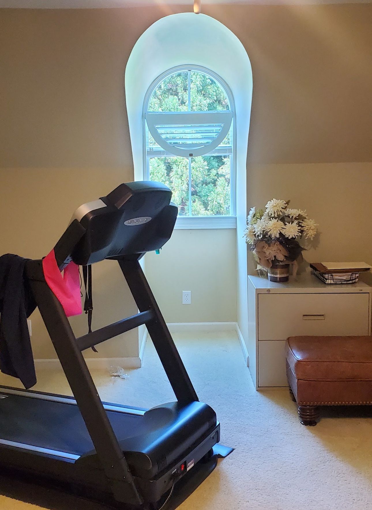 Treadmill in a room with a tall, arched window. A white cabinet and ottoman are nearby. Beige walls and carpet.