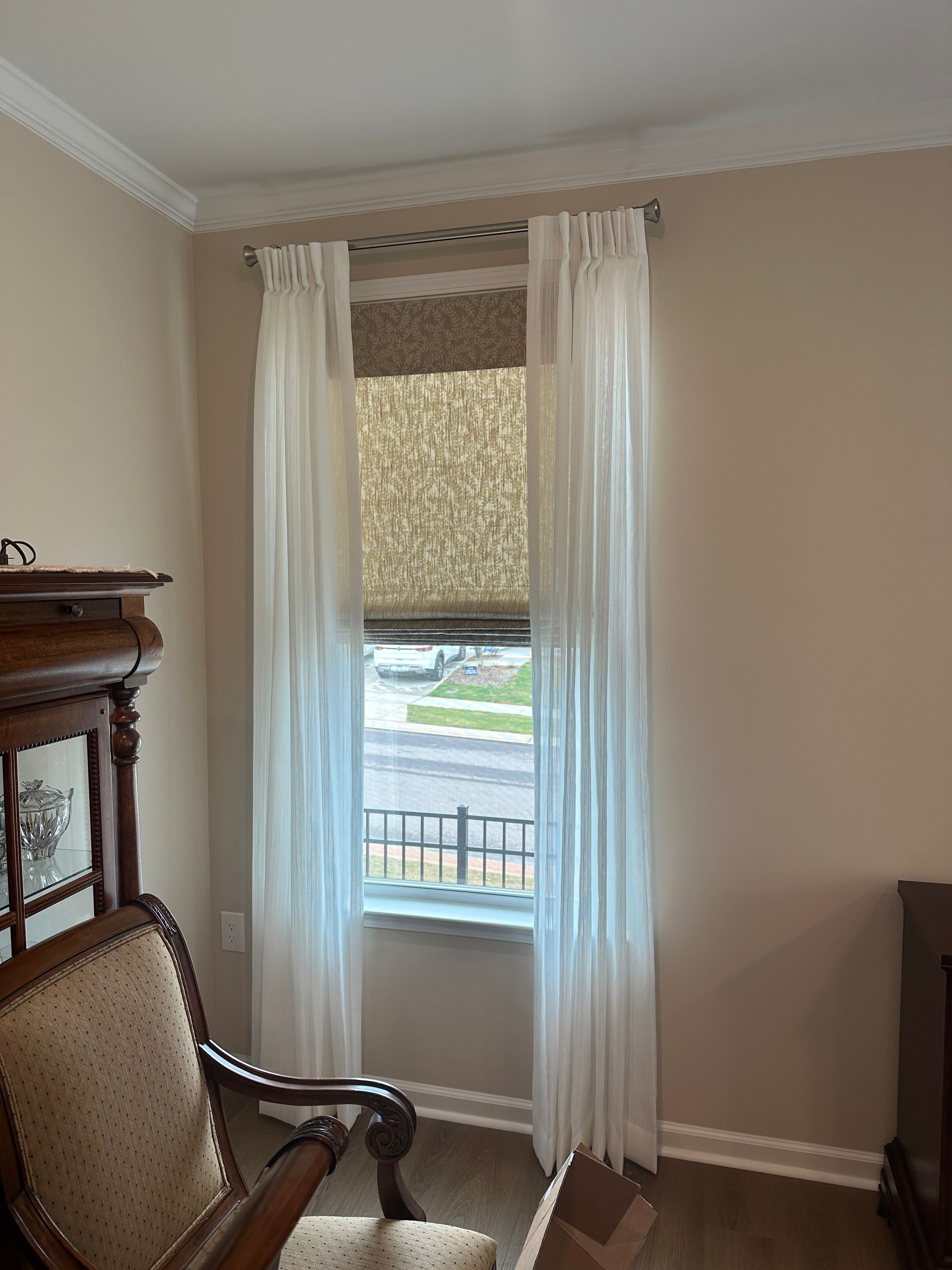 A window with sheer white curtains and a patterned shade, in a room with beige walls and wood furniture.