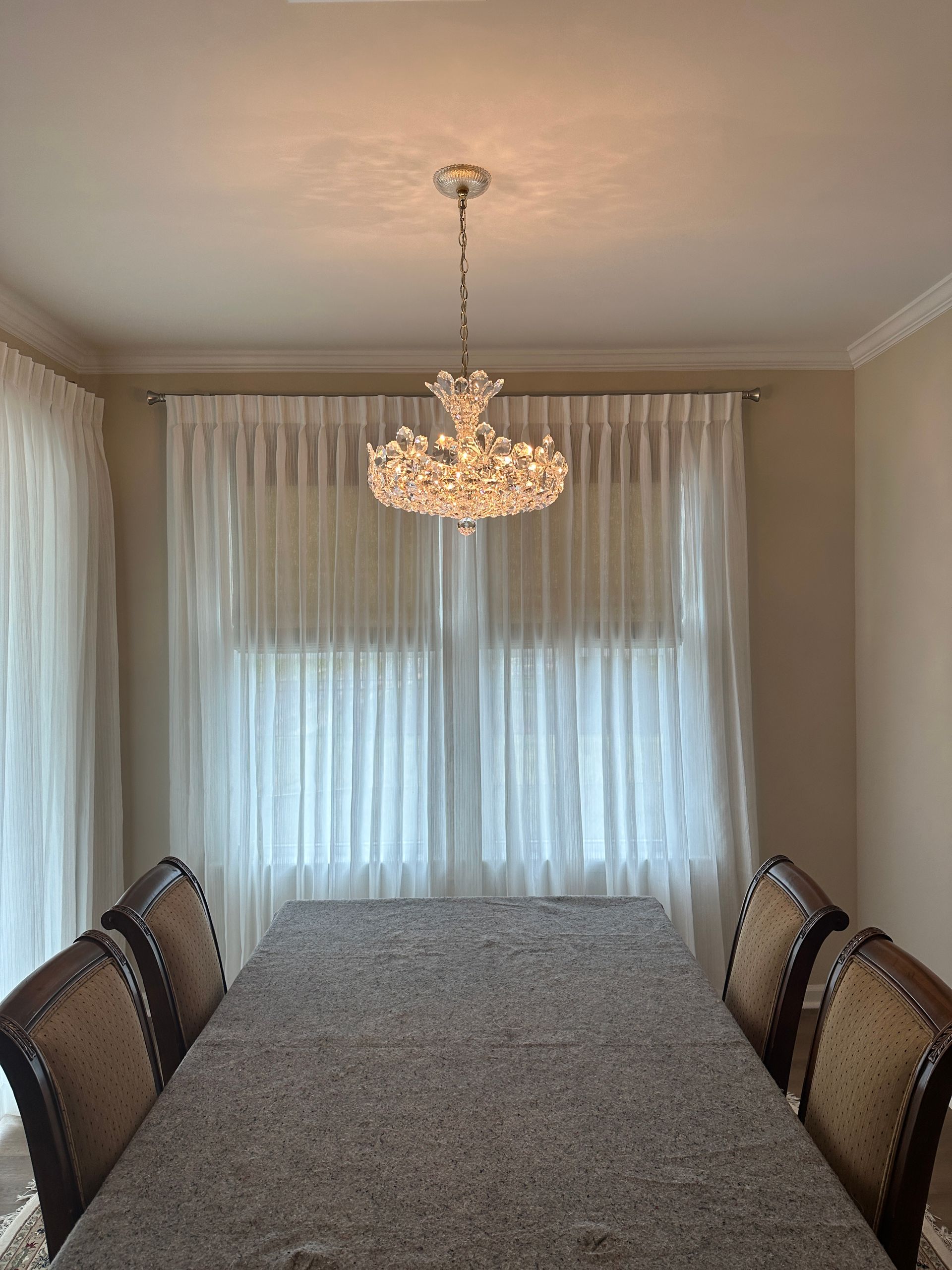Dining room with chandelier, long table, six chairs, and sheer white curtains.