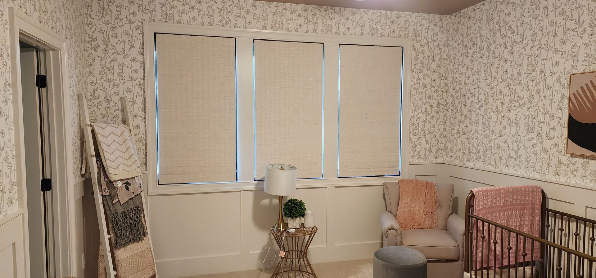 A nursery with floral wallpaper, three windows with blinds, a crib, and a chair with a pink blanket.