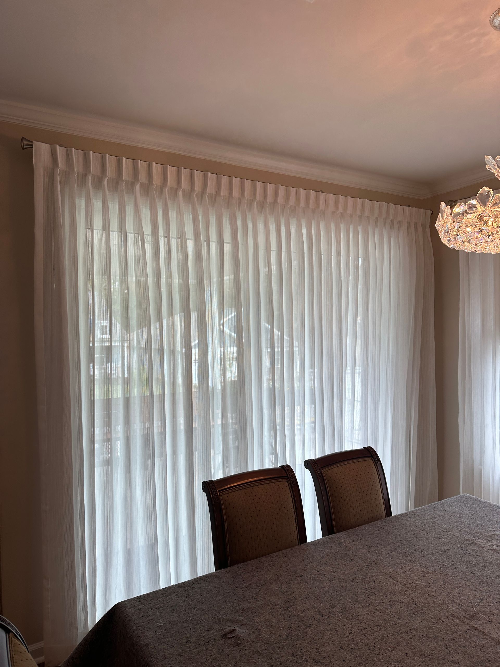 White sheer curtains cover a window, with two brown chairs and part of a table in the foreground.
