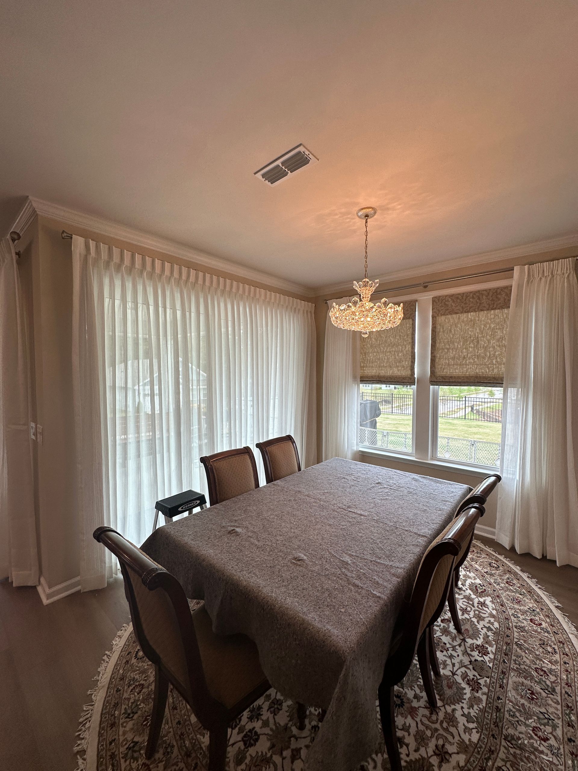 Dining room with long white curtains, chandelier, rectangular table with chairs, and patterned rug.