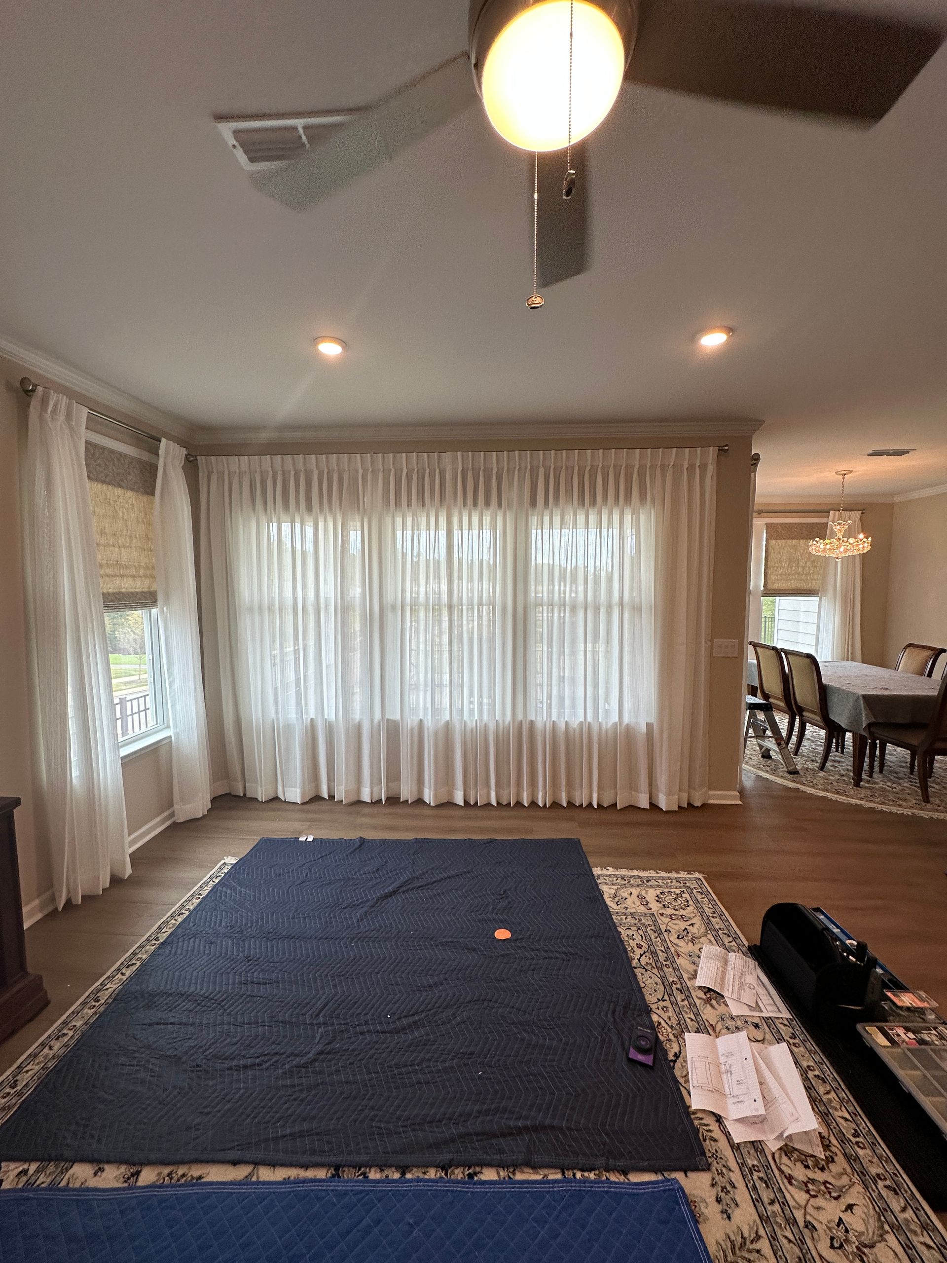 A living room with sheer curtains, a rug, a ceiling fan, and a dining room visible in the background.