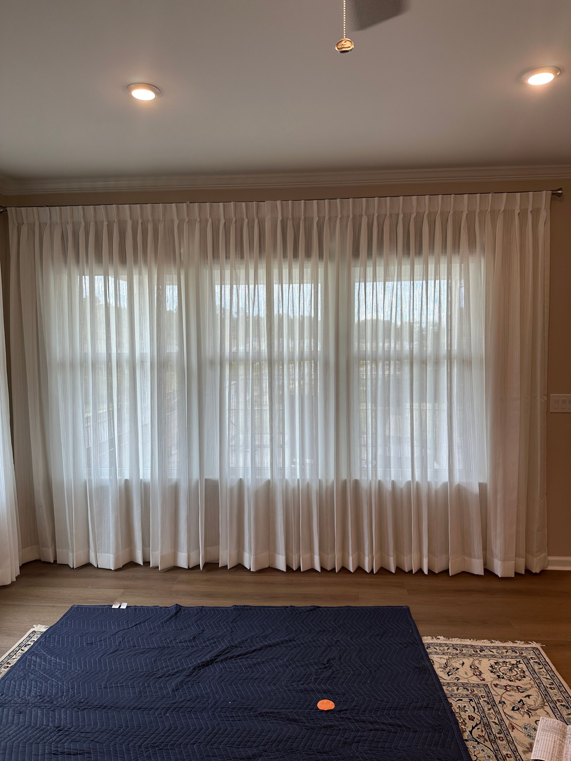 White sheer curtains covering a three-panel window. A blue rug sits on the wooden floor.