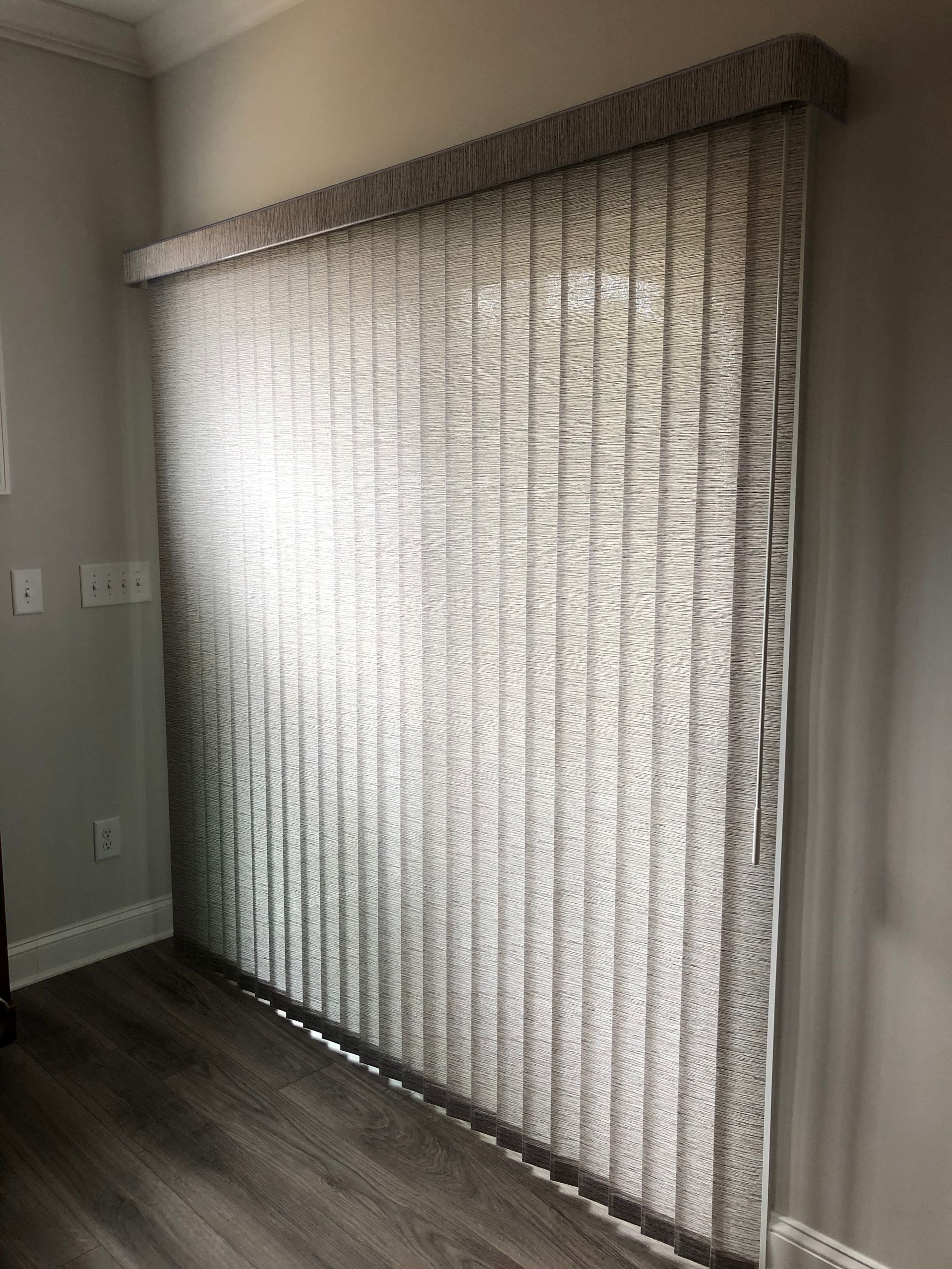Gray vertical blinds covering a large window, next to a wood floor and gray wall.