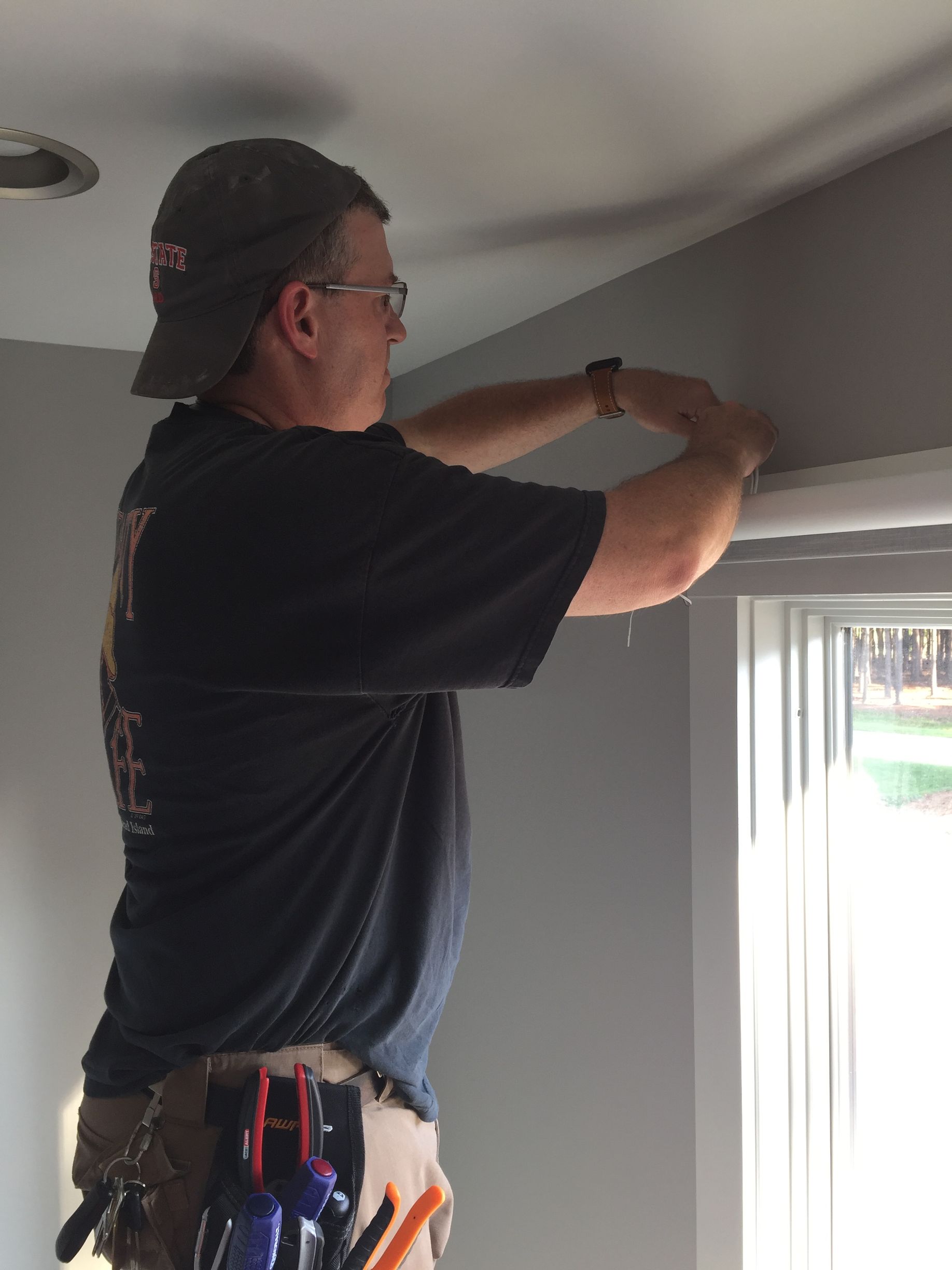 Person installing blinds near a window, wearing a cap and tool belt.