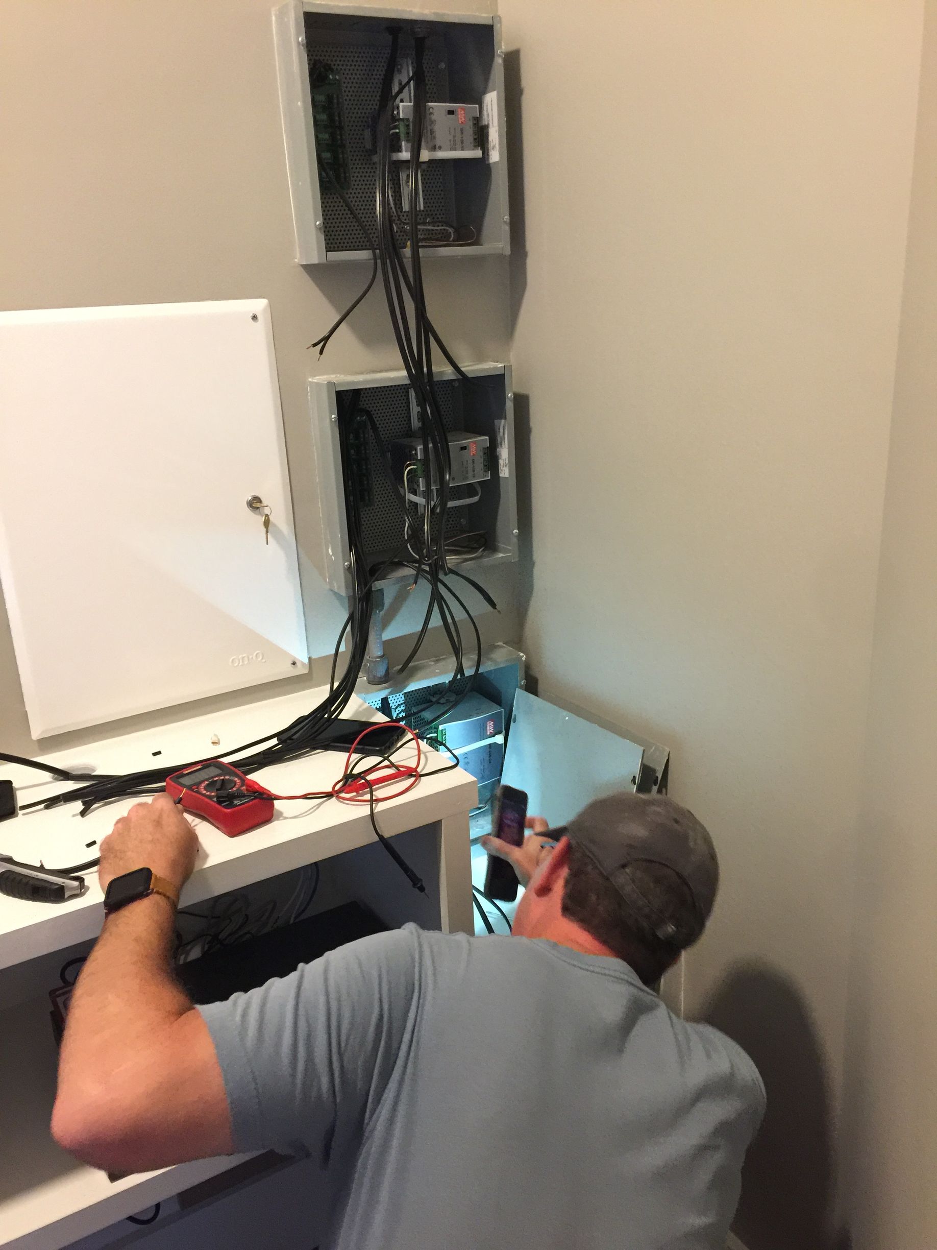 Man working on electrical equipment near wall-mounted panels. Wires and tools are visible; neutral color walls.