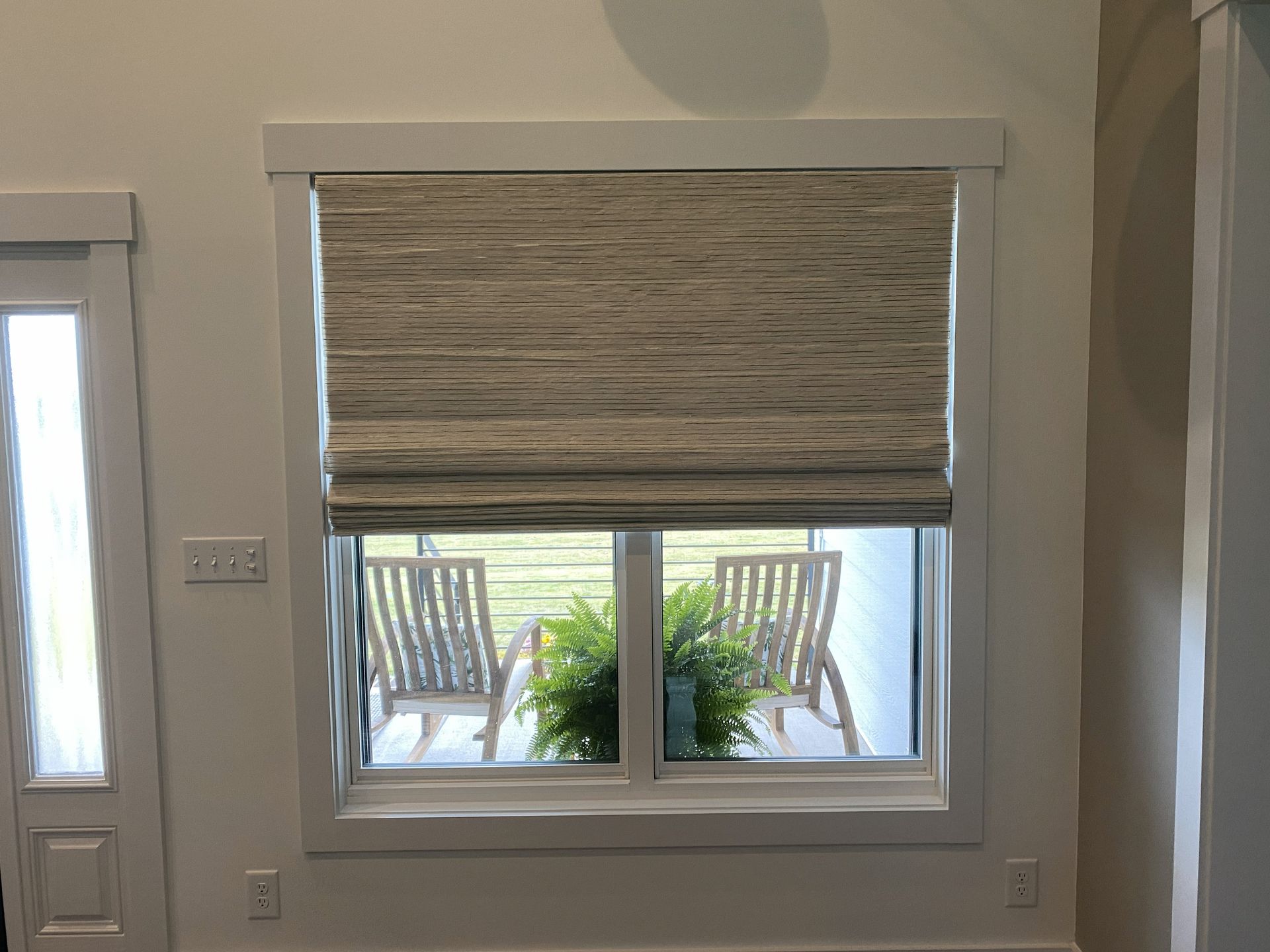 Window with woven shade, white frame. View of chairs and foliage outside.