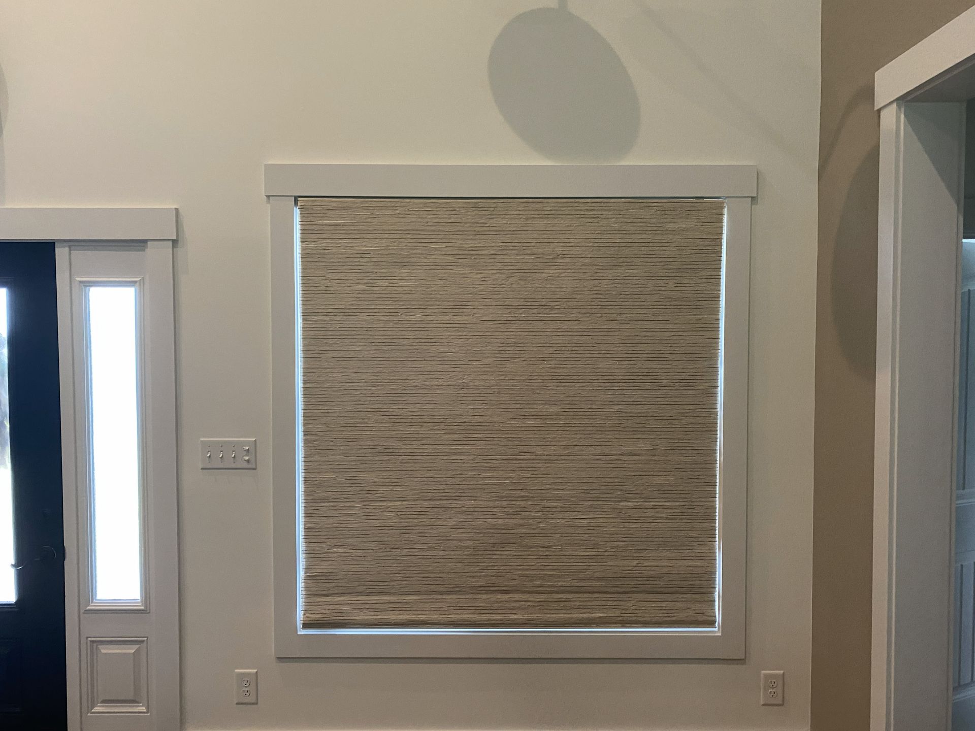 Tan woven shade inside a white-framed window on a beige wall.  A partially visible door is to the left.