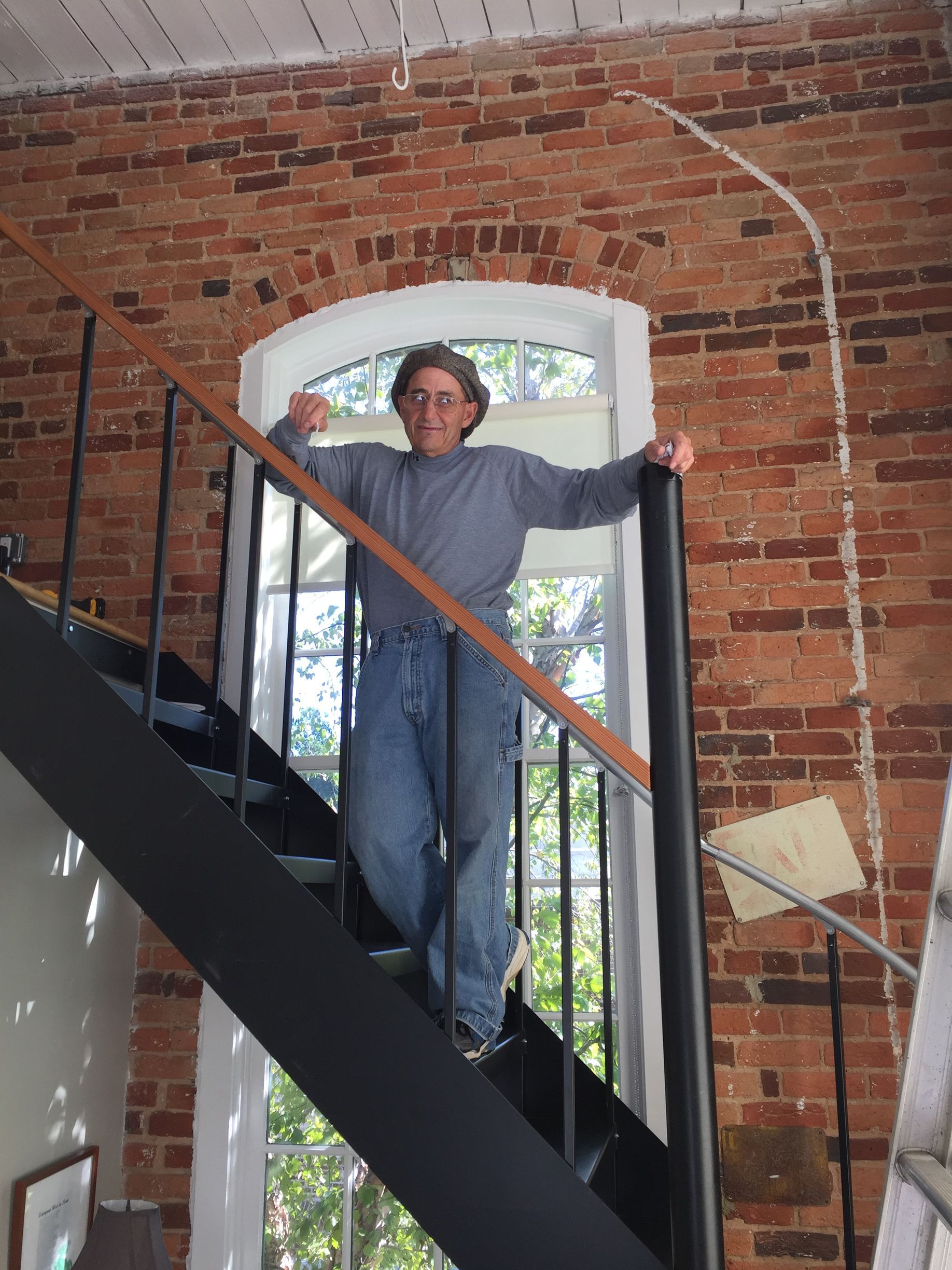 Person standing on black spiral staircase in front of arched window, brick wall.