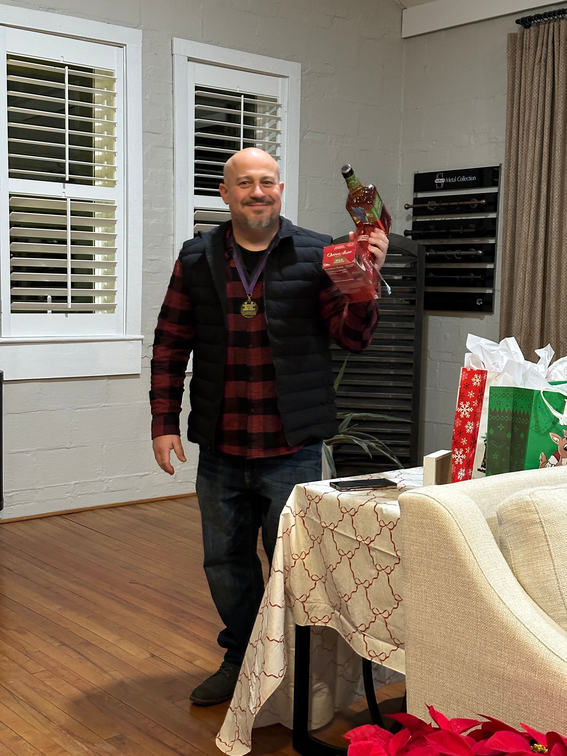 Man holding a trophy, smiling. Wearing a red plaid shirt, black vest, and jeans indoors.