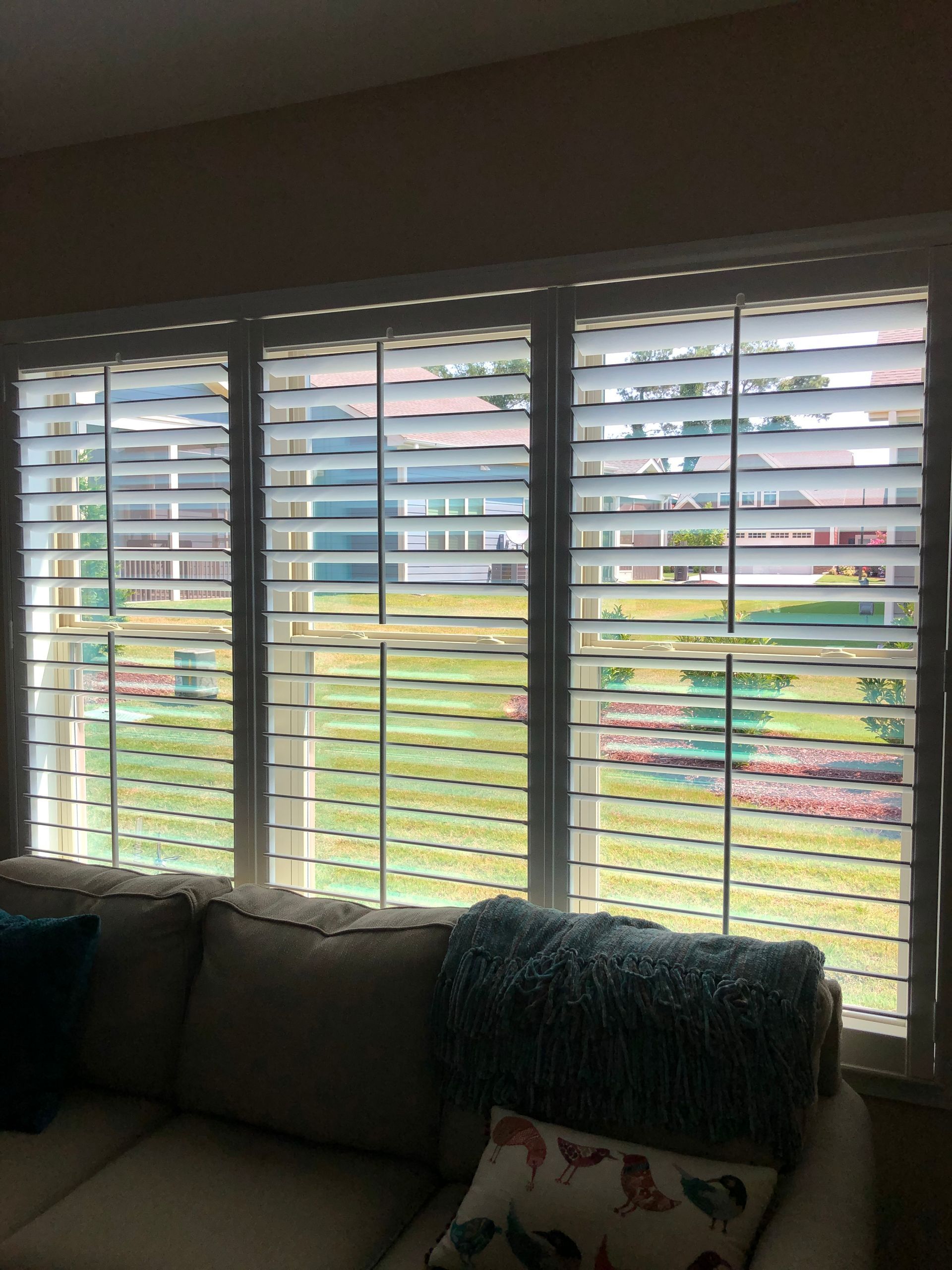 White window shutters over a couch with pillows, looking out to a garden with green grass and flowers.