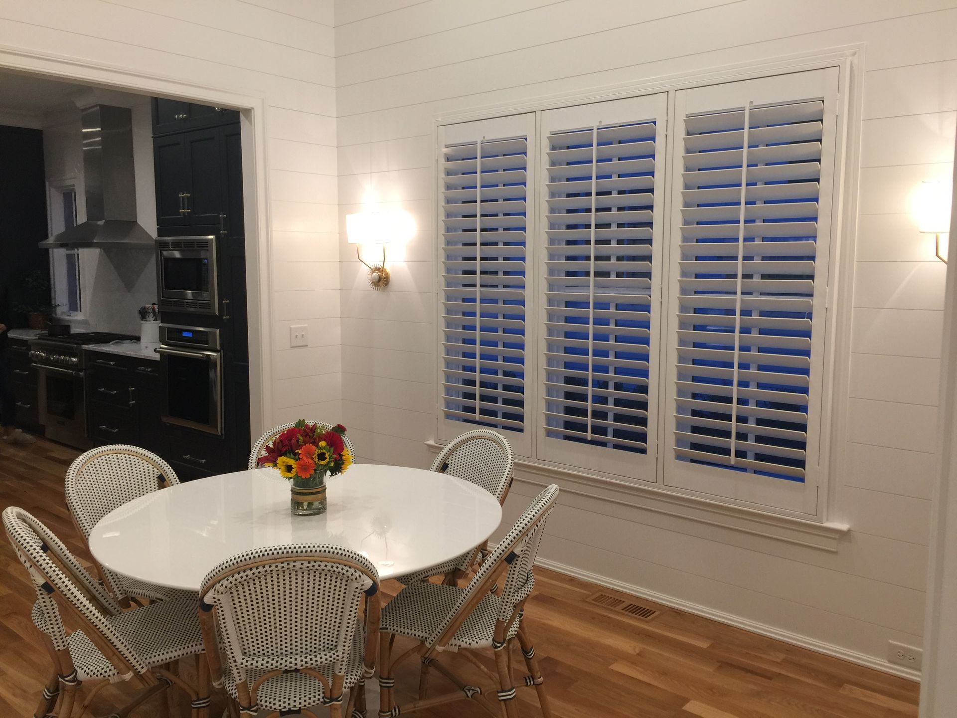Dining room with white shutters, round table, woven chairs, and kitchen view.