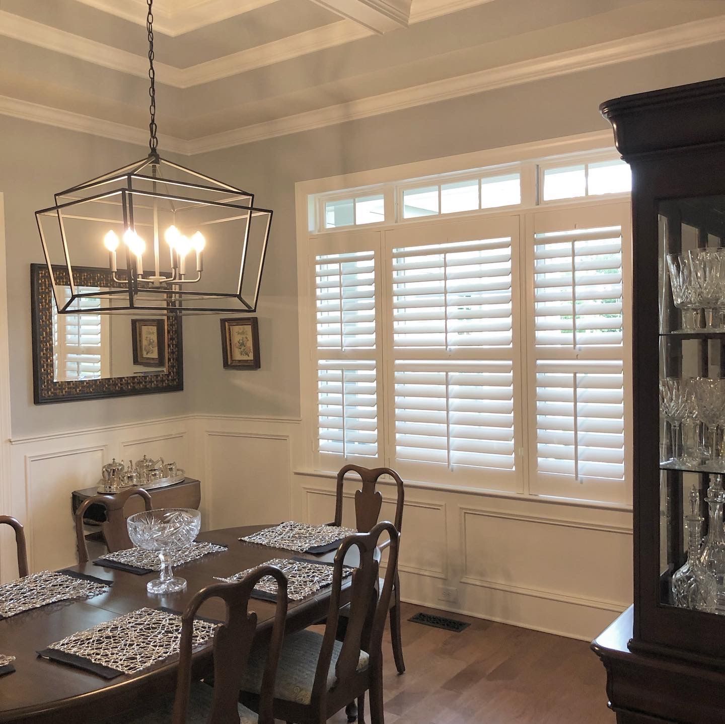 Dining room with table set for guests, chandelier, white shutters, and a dark wood hutch.