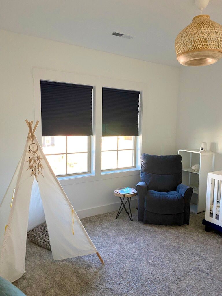 A light-filled nursery with a teepee, dark blue window shades, a rocking chair, and a crib.