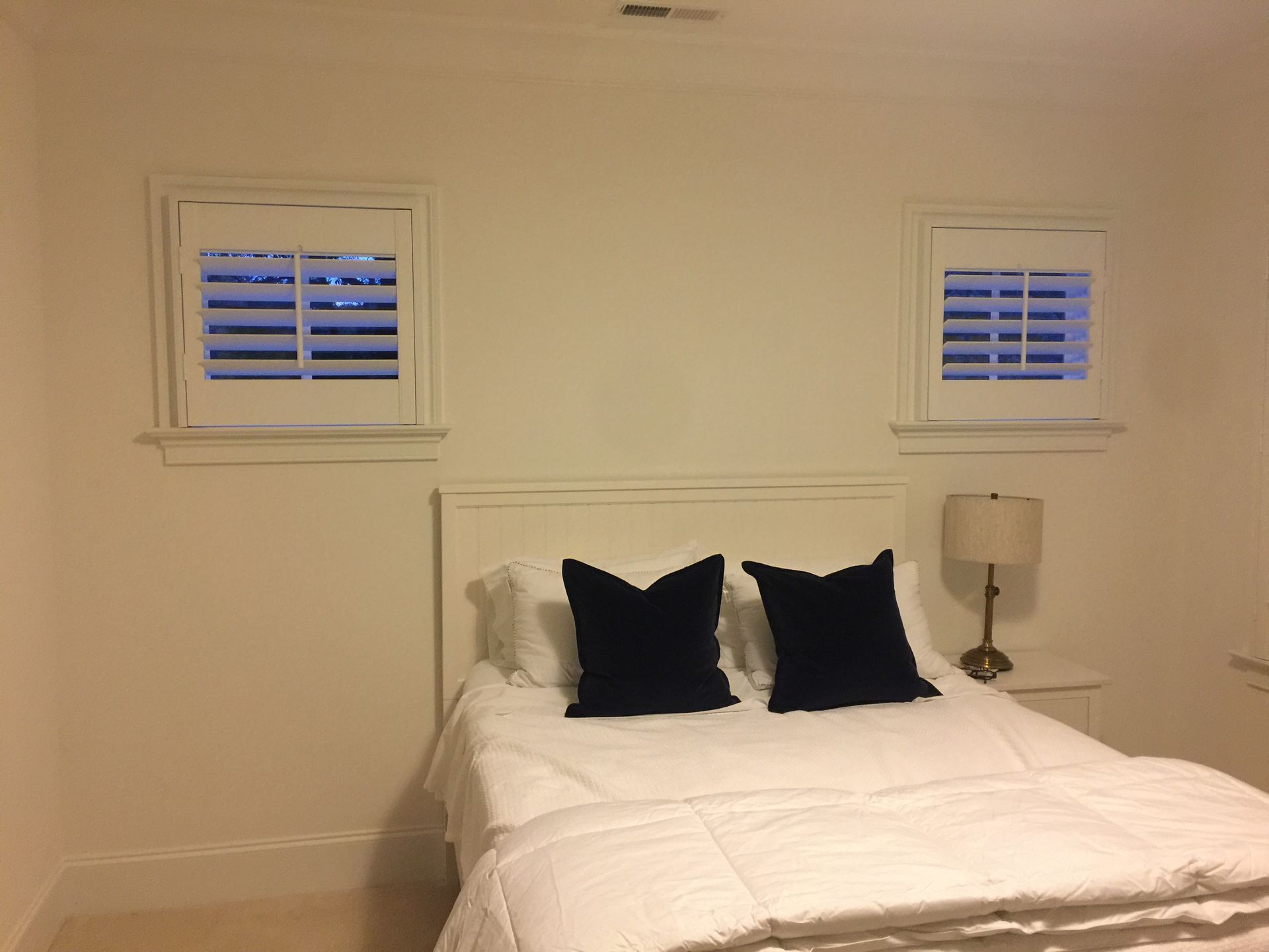 Bedroom with white walls, bed, two small windows with blinds, and a lamp on a nightstand.