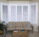 White shutters on a bay window above a tan couch and wicker coffee table.