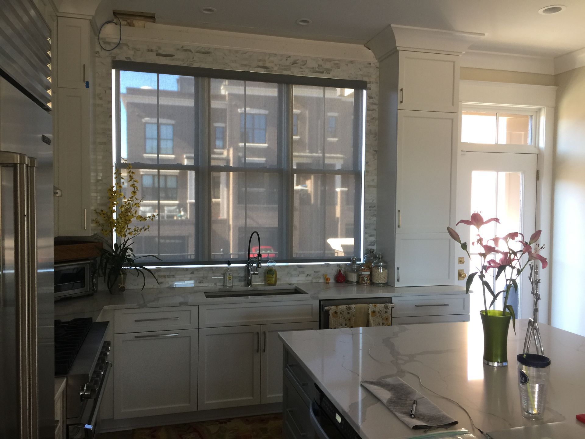 Bright kitchen with white cabinets, marble counters, and a large window with a roller shade.