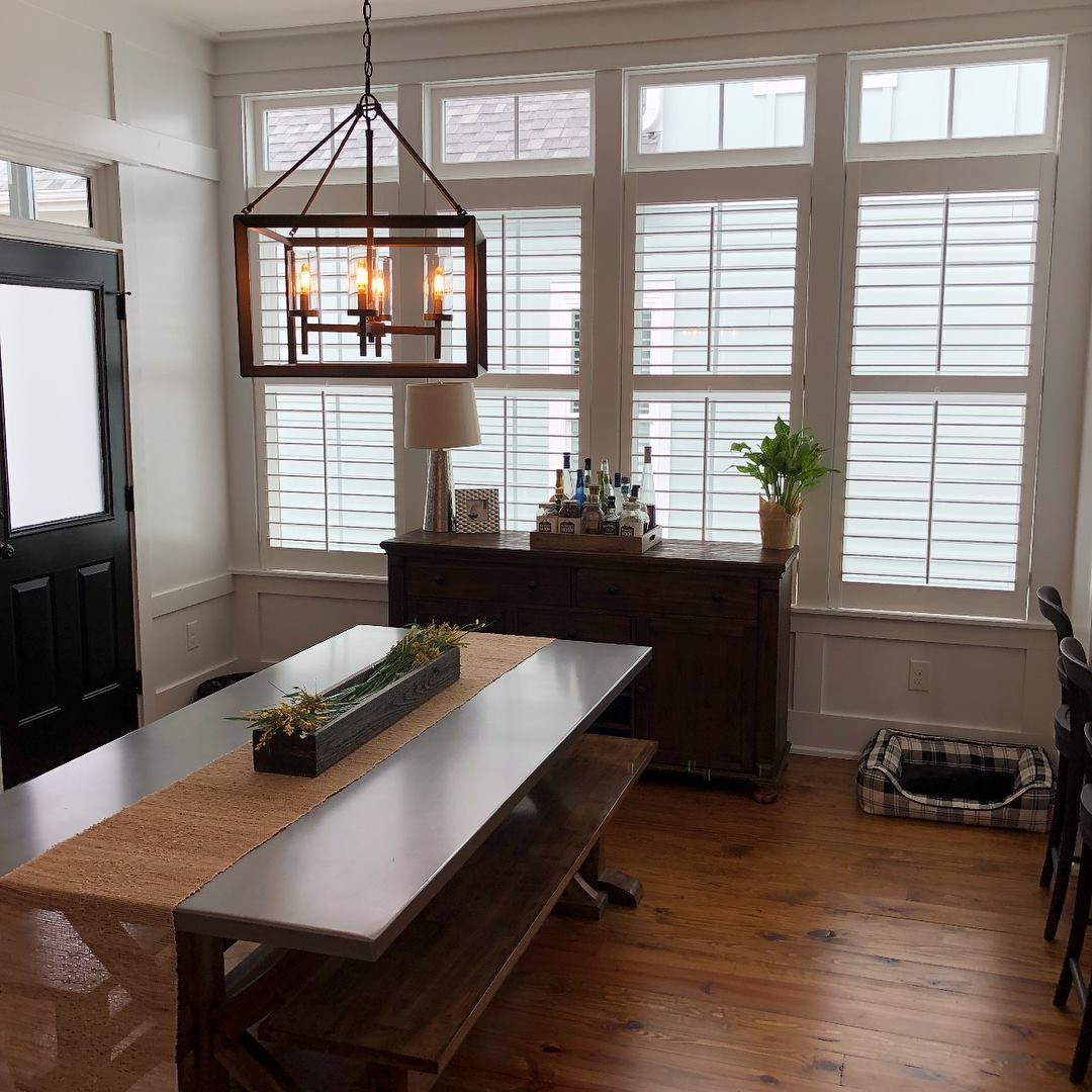 Dining room with a long table, benches, and a wooden buffet. Windows with shutters and a chandelier.