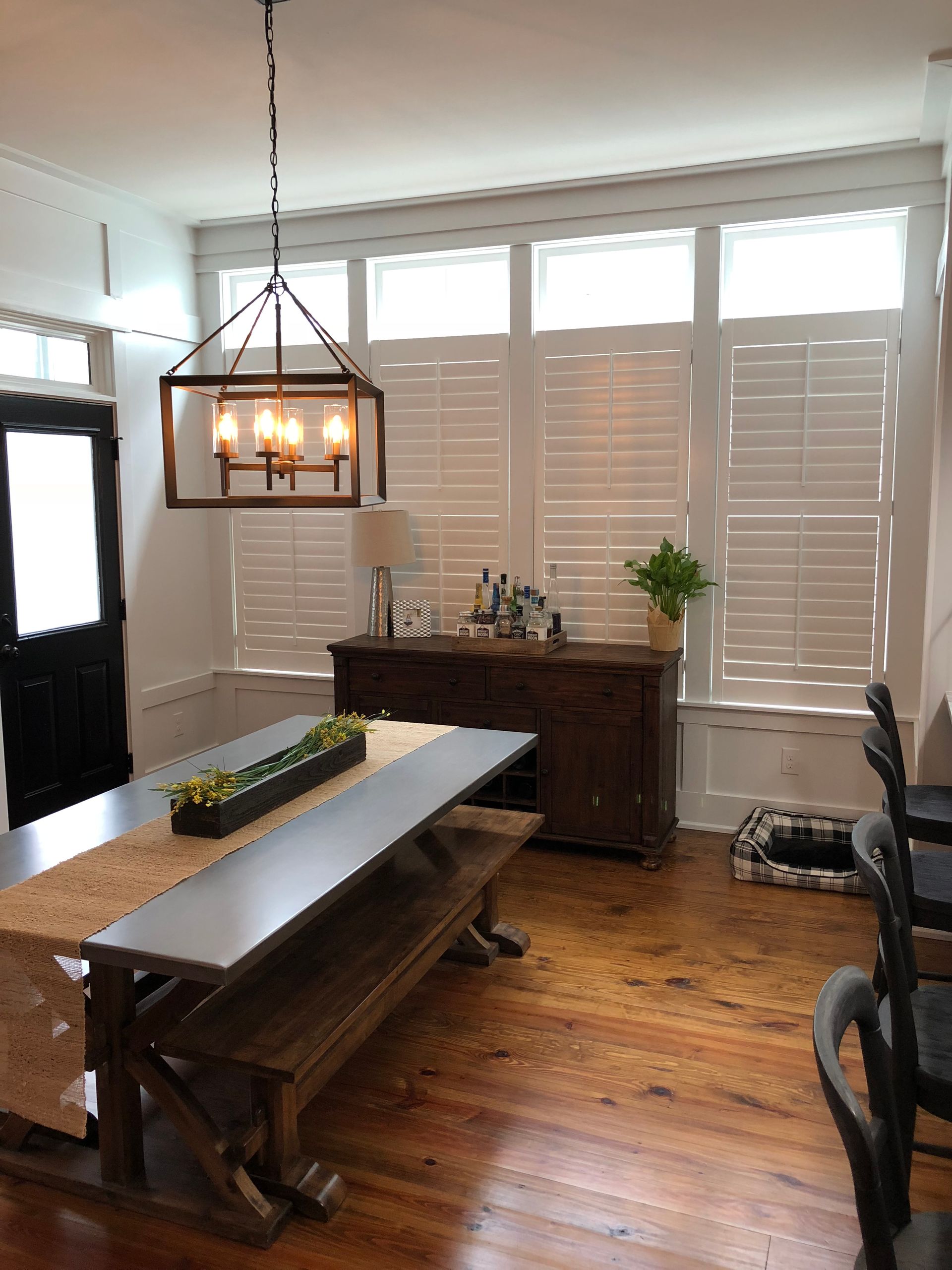 Dining room with wooden table, benches, and a dark wooden buffet. Windows with white shutters.