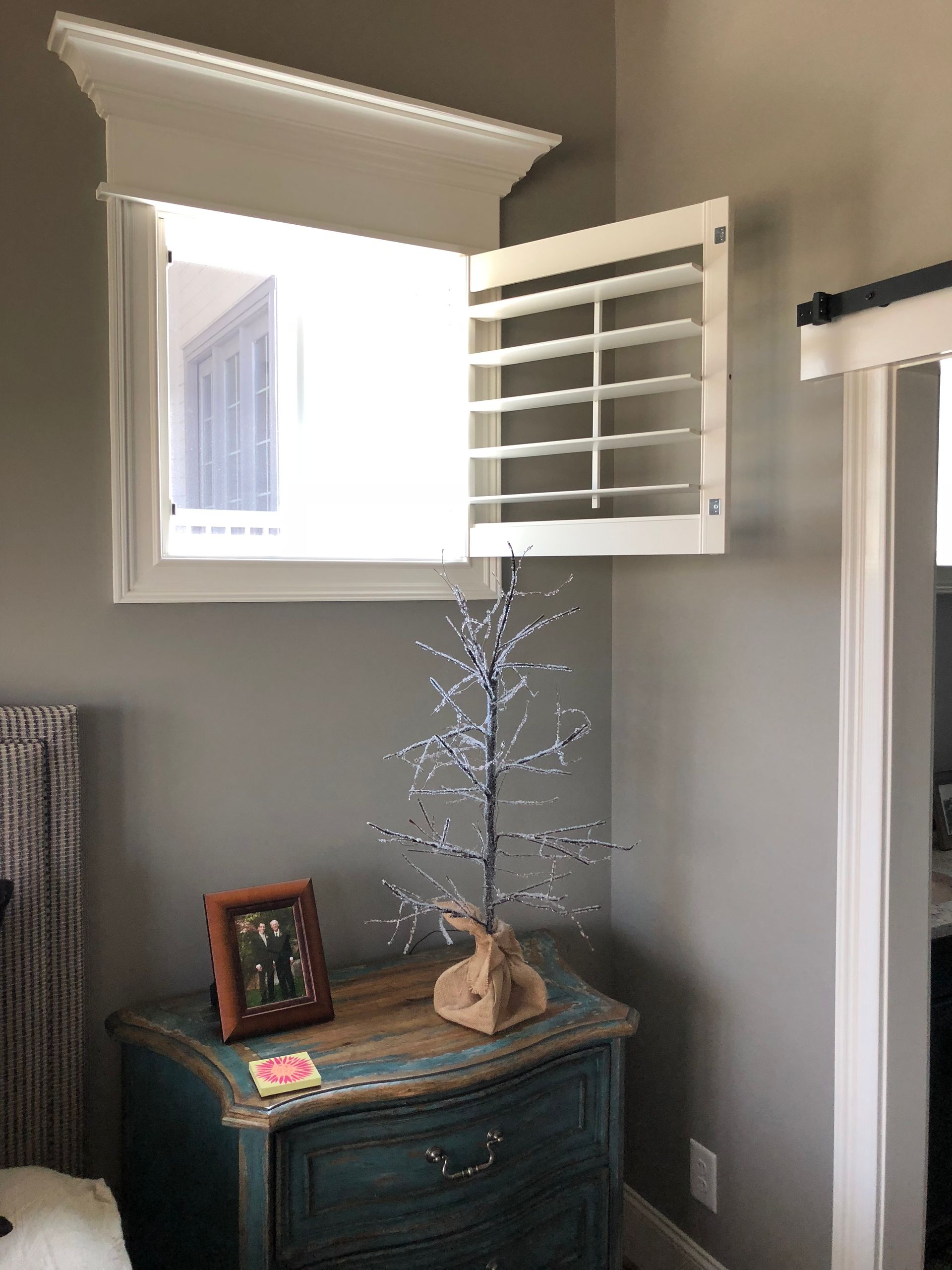 White shutters open on a bedroom window, above a teal nightstand with a small tree and framed photo.