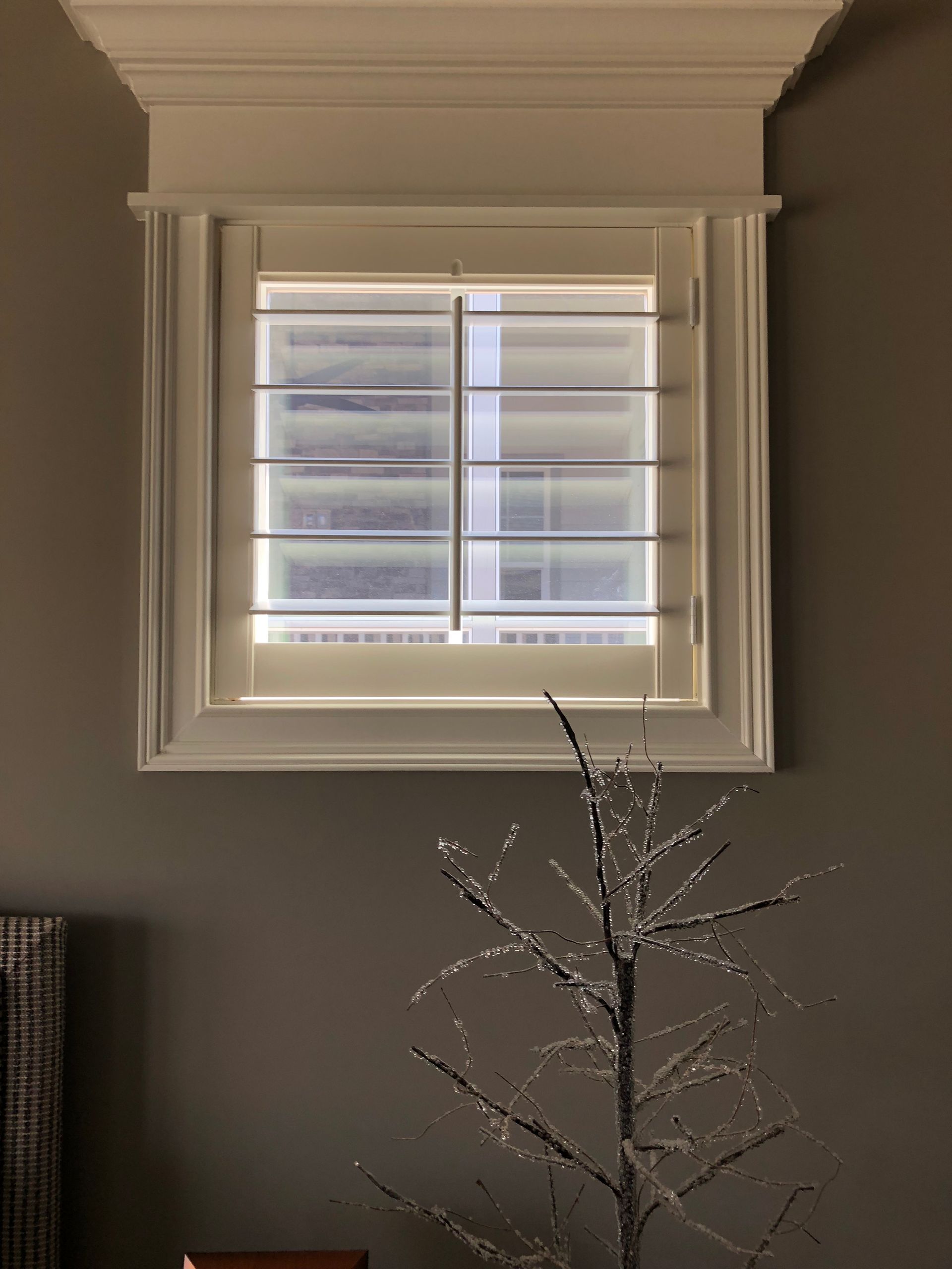 White framed window with horizontal shutters, under a decorative molding, on a gray wall.