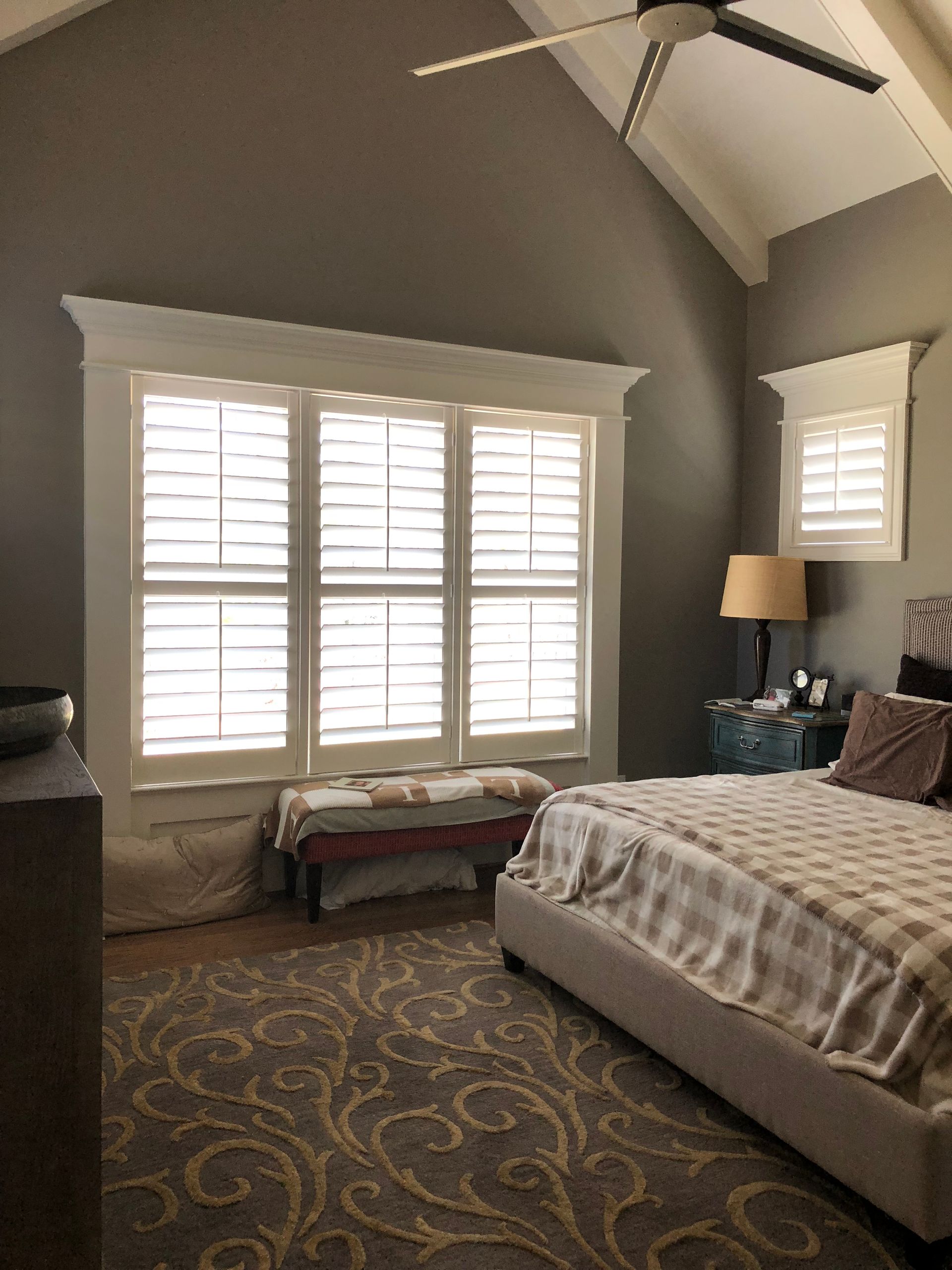 Bedroom with gray walls, white shutters, a bed with a cream blanket, and patterned rug.