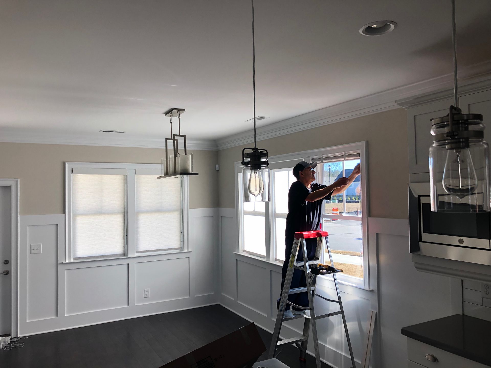 Man on a ladder installing window blinds in a dining room with white trim, tan walls, and a dark floor.