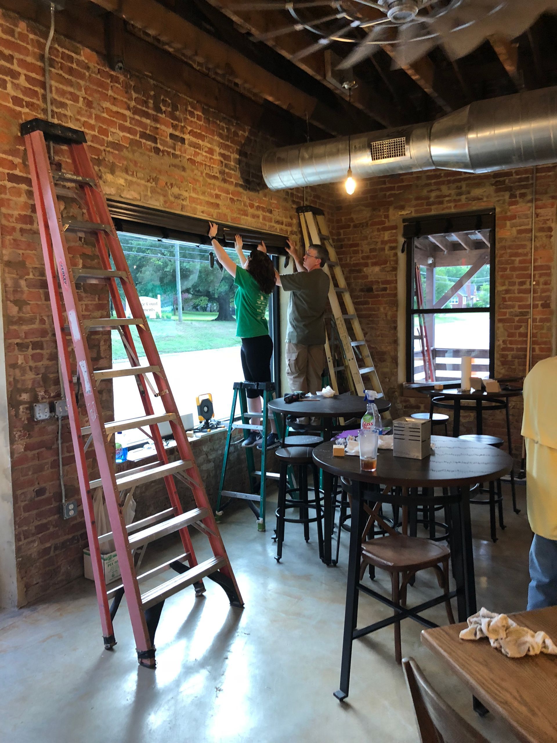 People installing a window in a brick-walled room using ladders. Circular tables and chairs are nearby.