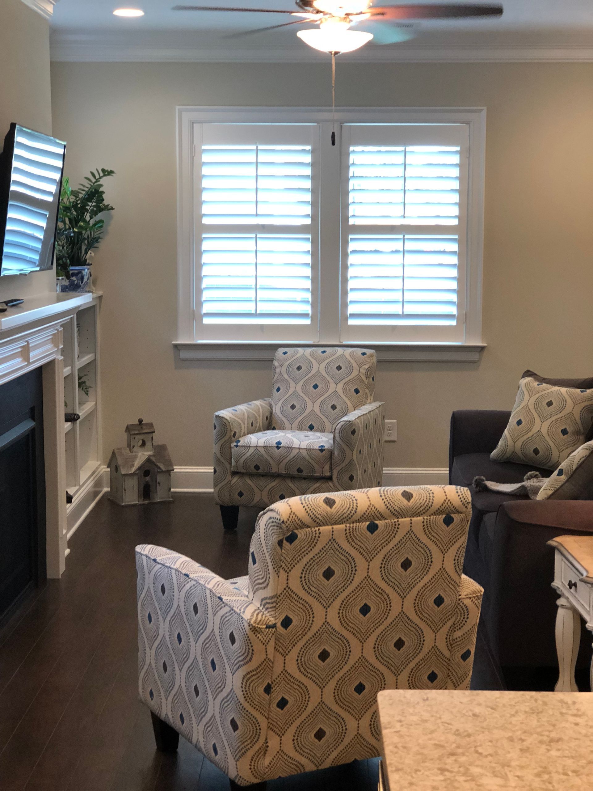 Living room with patterned armchairs, dark sofa, window shutters, and fireplace.