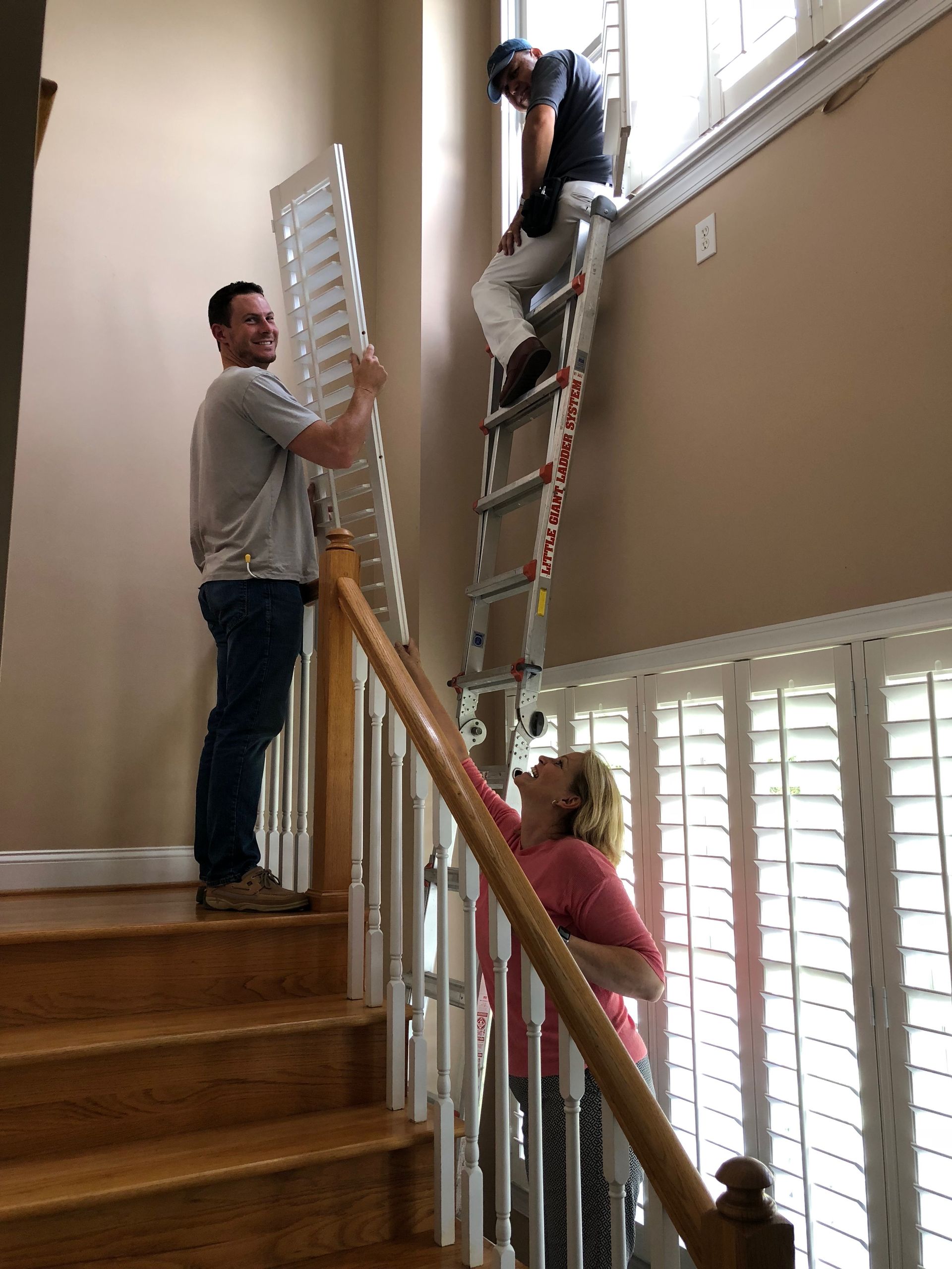 Three people installing shutters on a tall window. One on a ladder, two others assisting. Staircase visible.