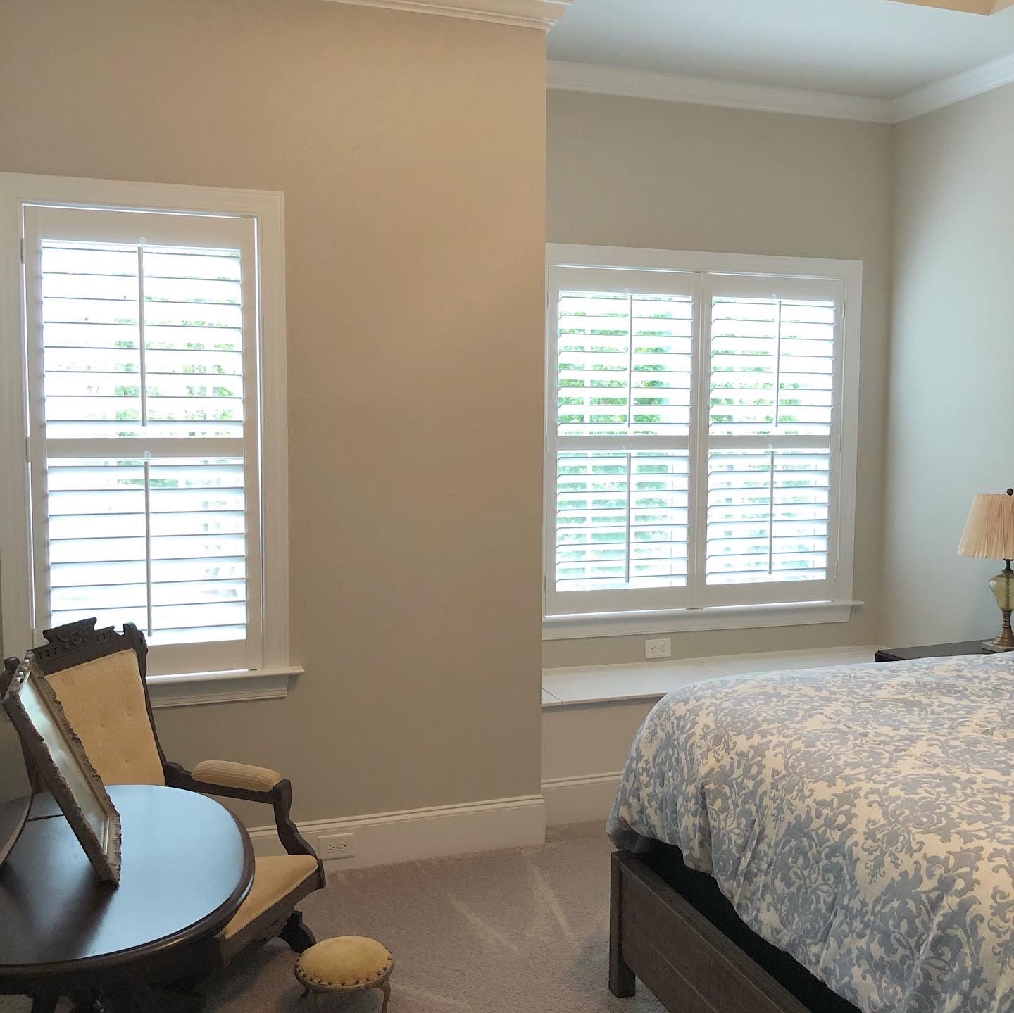 Bedroom with white-framed windows, shutters, a round table, ornate chair, bed with patterned cover.