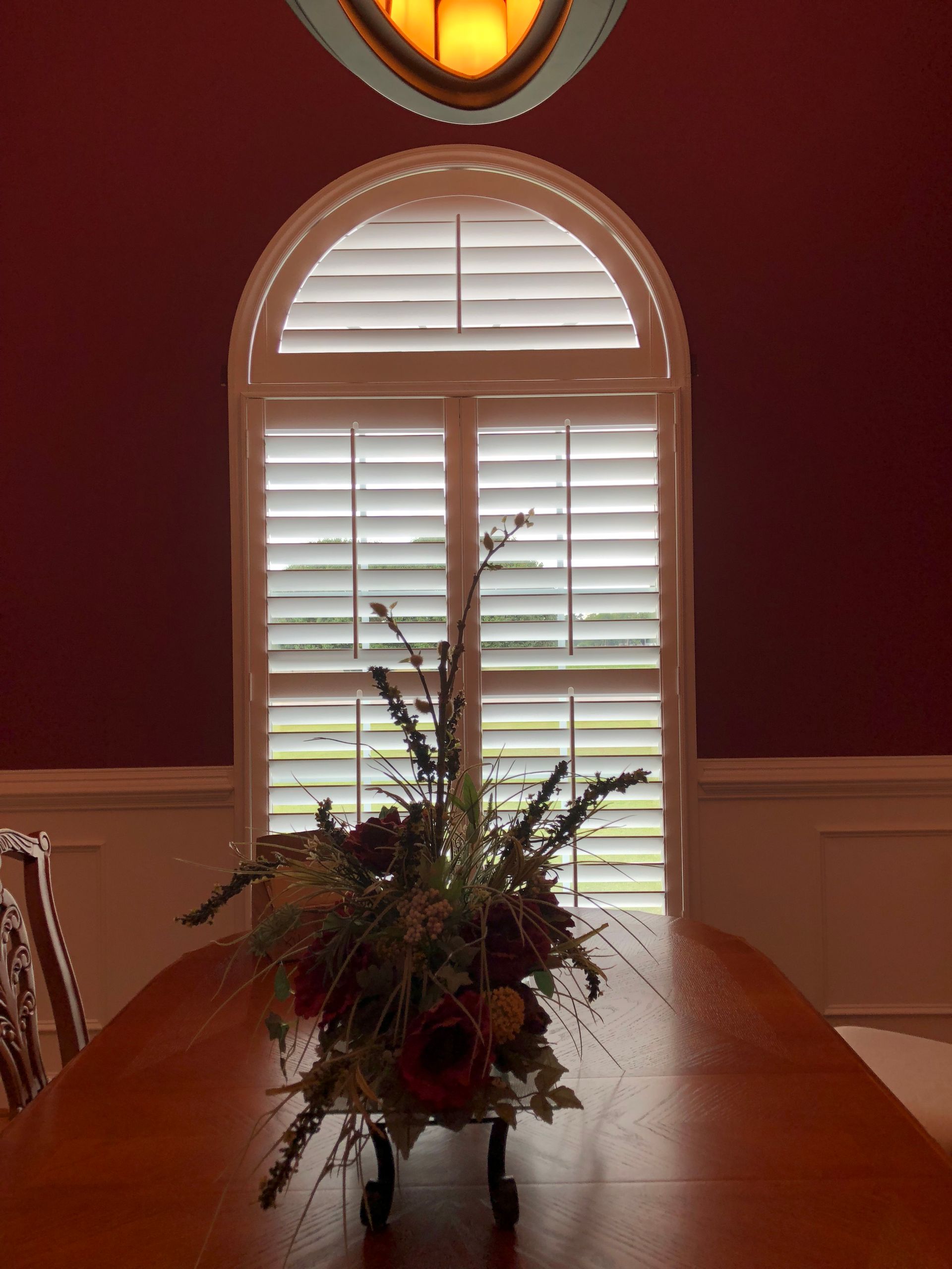 Dining room with table, centerpiece, and arched window with shutters. Dark red walls.