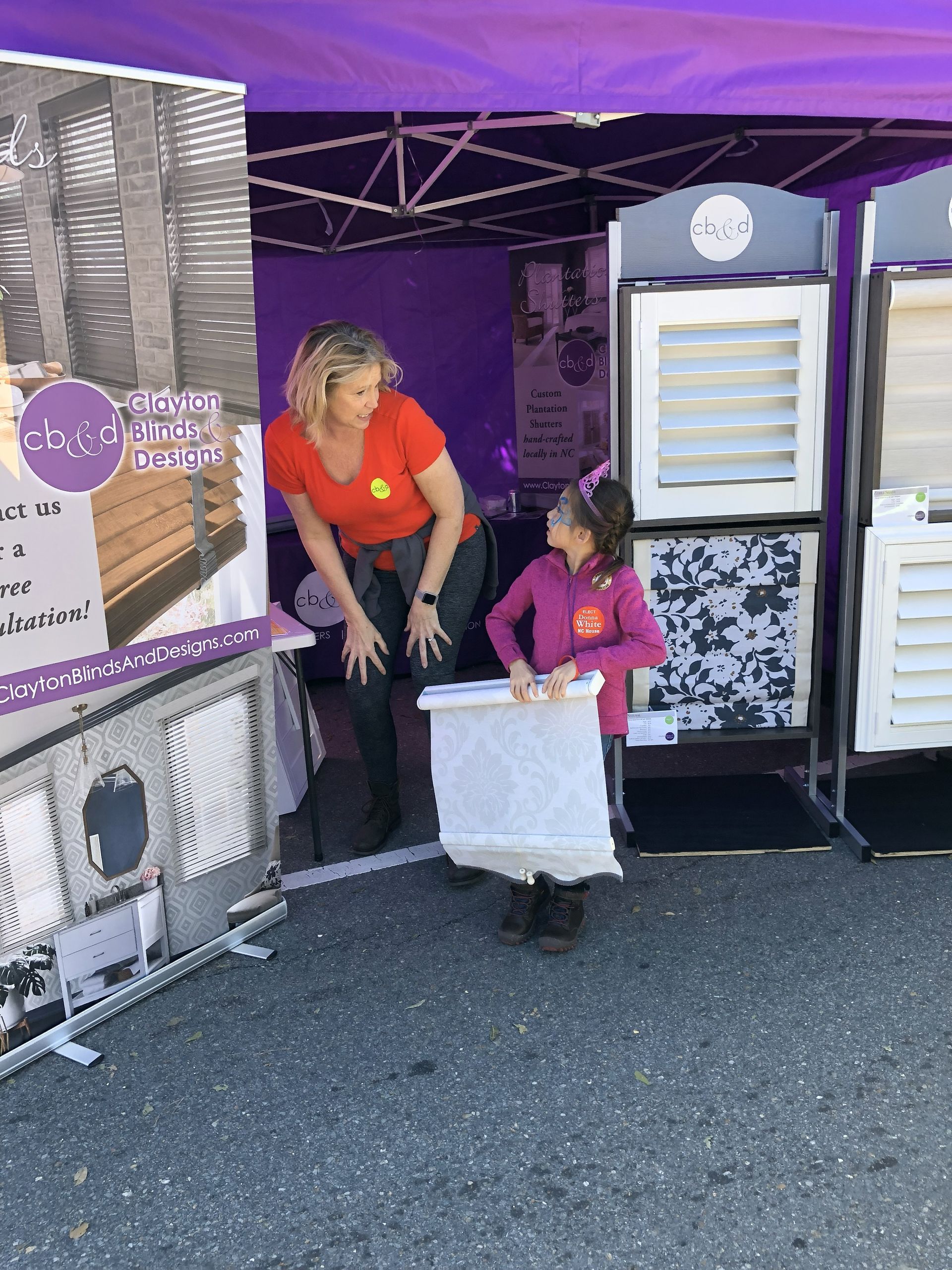 Woman in orange shirt with child holding a white display. They stand in front of a booth with window shutter samples.