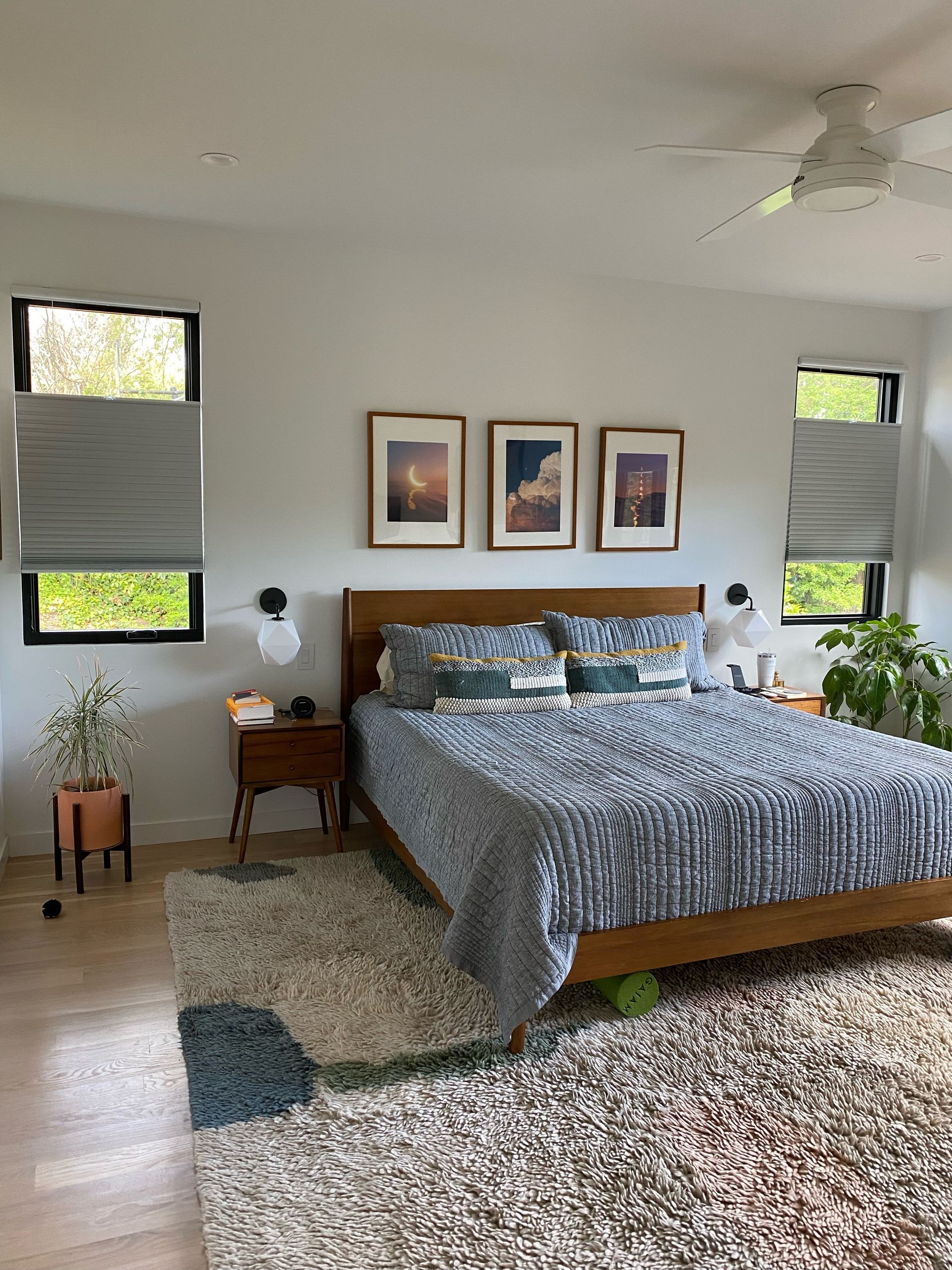 Bedroom with bed, art, windows, rug, and plants. Neutral walls, wood furniture, blue and brown decor.
