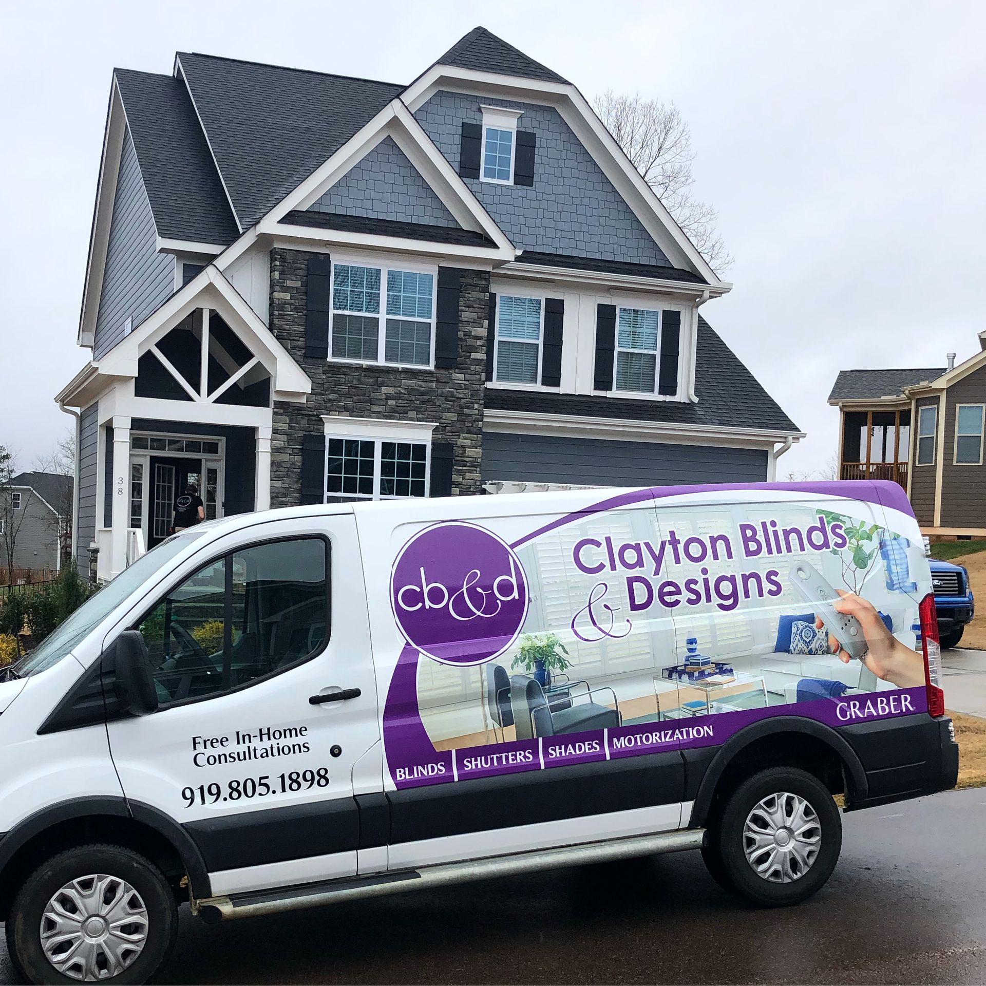 A Clayton Blinds Designs van parked in front of a house. The van has the company logo in purple and white.