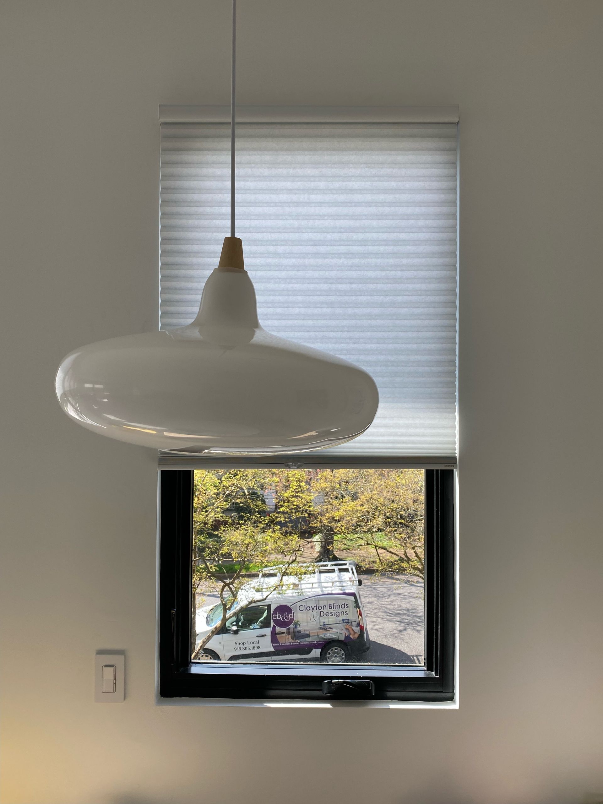 White pendant lamp hanging in front of a window with blinds and a view of trees and a truck.