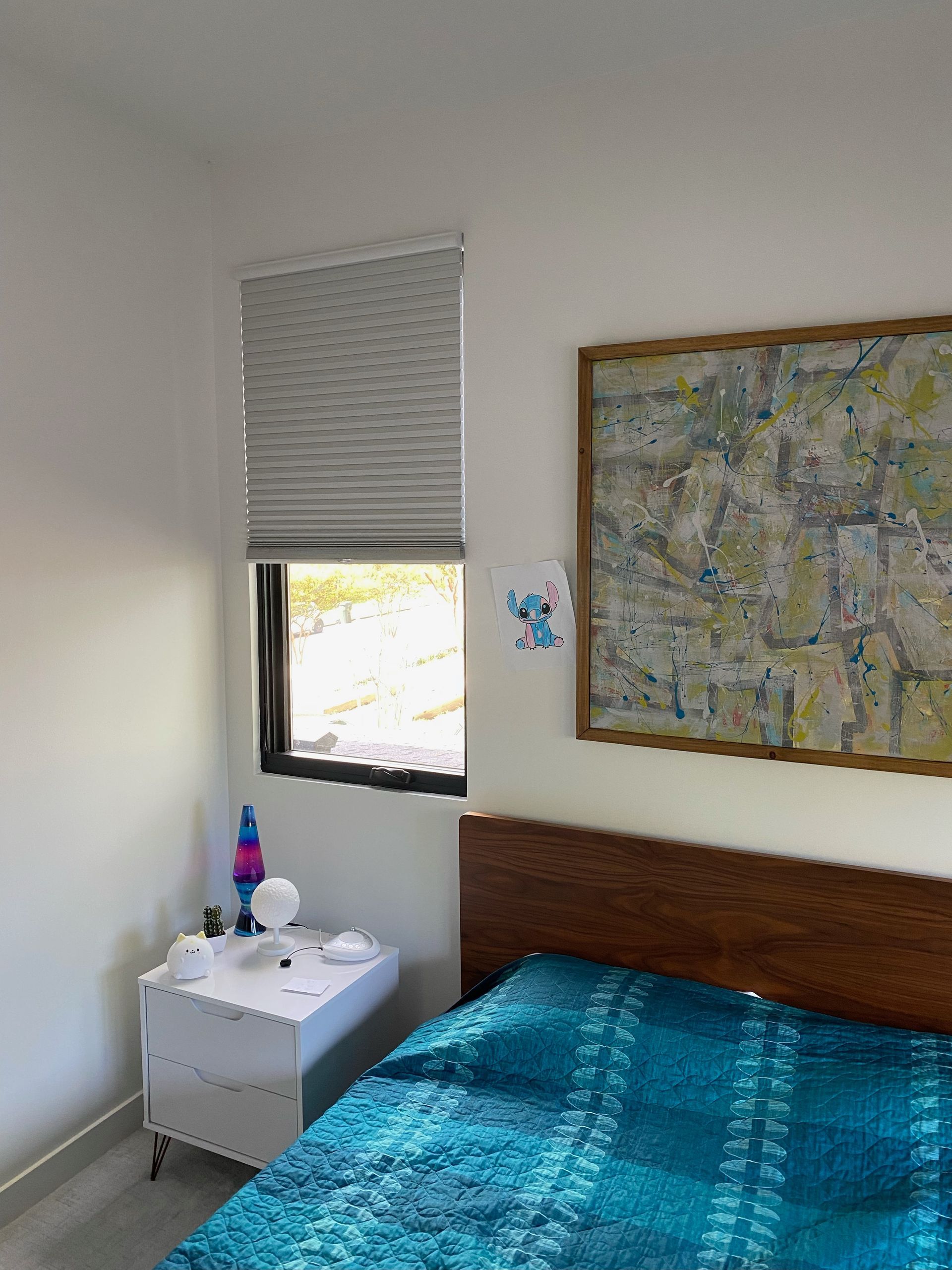 Bedroom corner: bed with blue quilt, nightstand with lamp, window with blind, and art on wall.